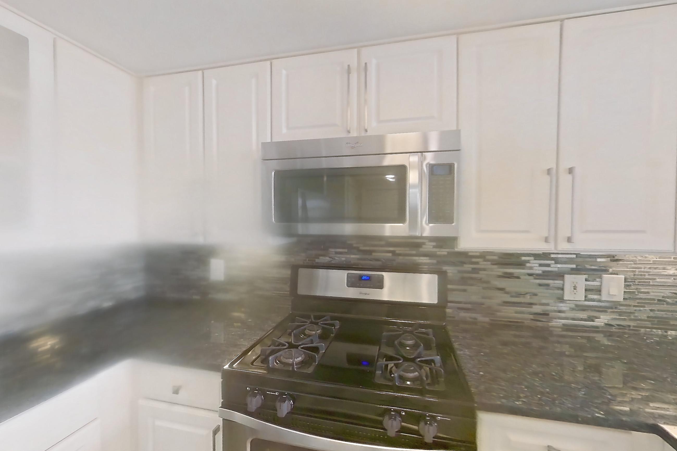 Kitchen with dark stone countertops, gas range, light countertops, light wood-finish flooring, microwave, oven/range, refrigerator, and stainless steel appliances, Apartment 911 at The Barton at Woodley