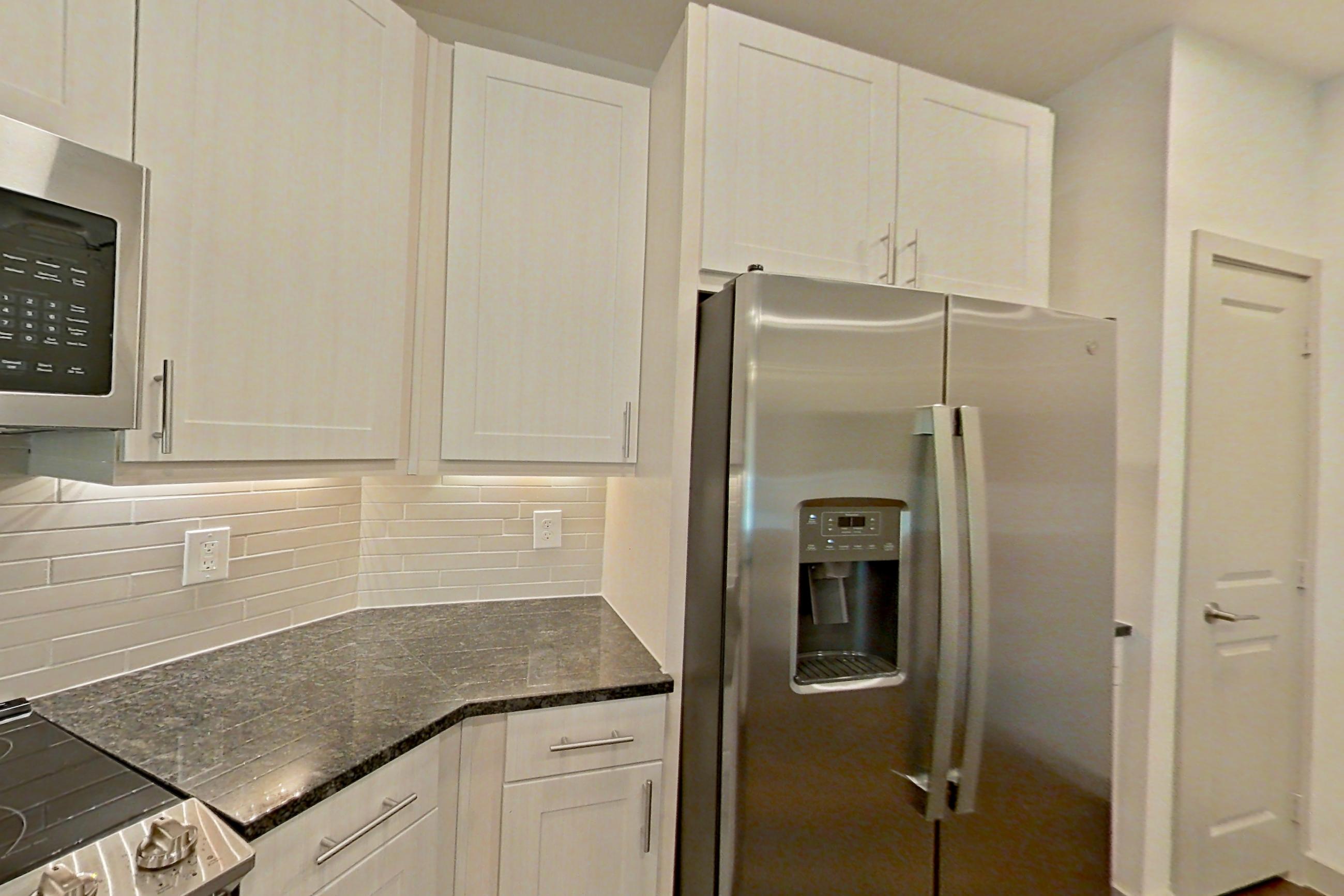 Kitchen with dark stone countertops, dark wood-finish flooring, dishwasher, electric range, light countertops, microwave, oven/range, refrigerator, stainless steel appliances, and water & ice dispenser, Apartment 1412 at Merit Lewisville