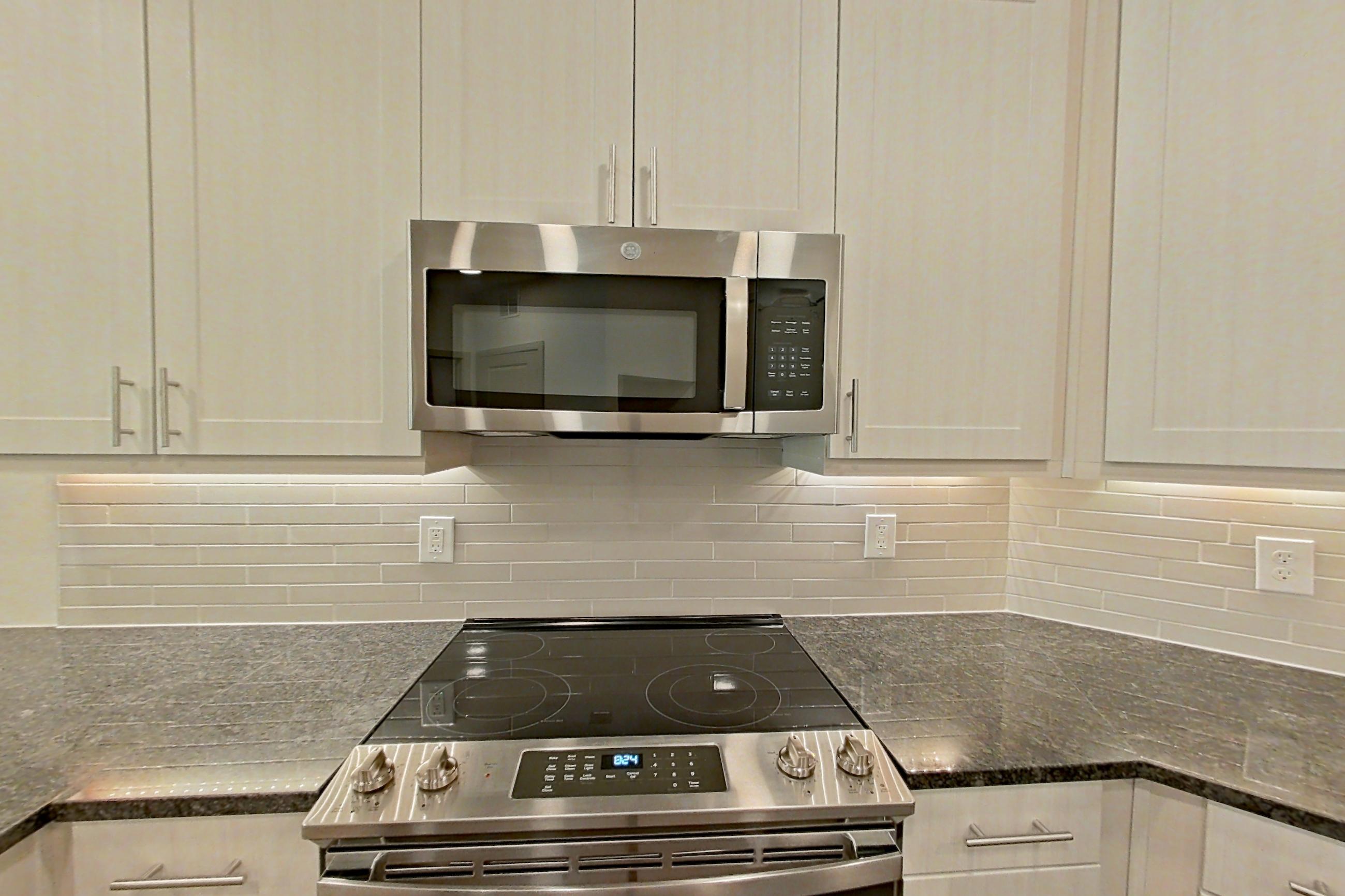 Kitchen with dark stone countertops, dark wood-finish flooring, dishwasher, electric range, light countertops, microwave, oven/range, refrigerator, stainless steel appliances, and water & ice dispenser, Apartment 1412 at Merit Lewisville