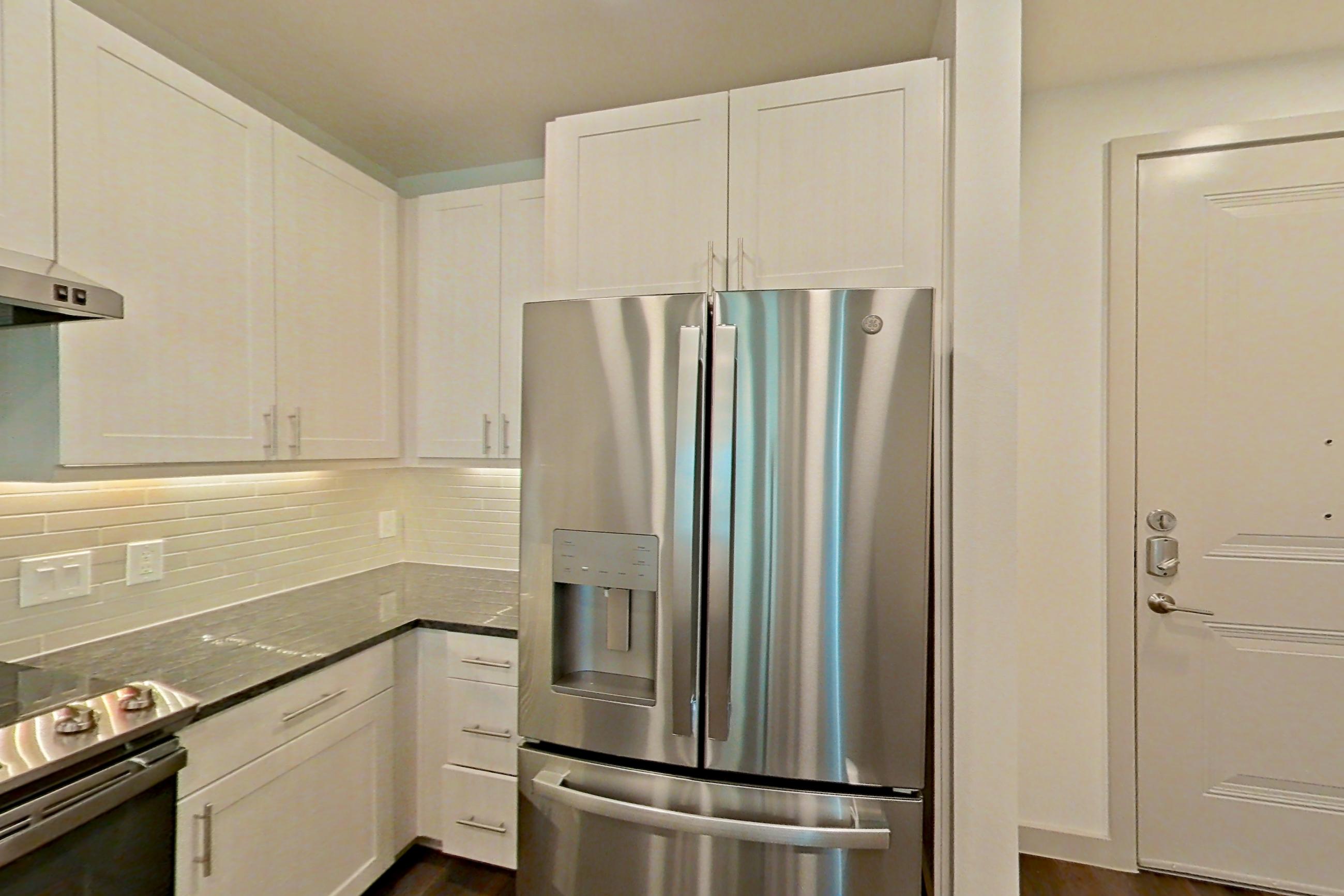 Kitchen with dark stone countertops, dark wood-finish flooring, dishwasher, electric range, oven/range, range hood, refrigerator, stainless steel appliances, and water & ice dispenser, Apartment 1208 at Merit Lewisville