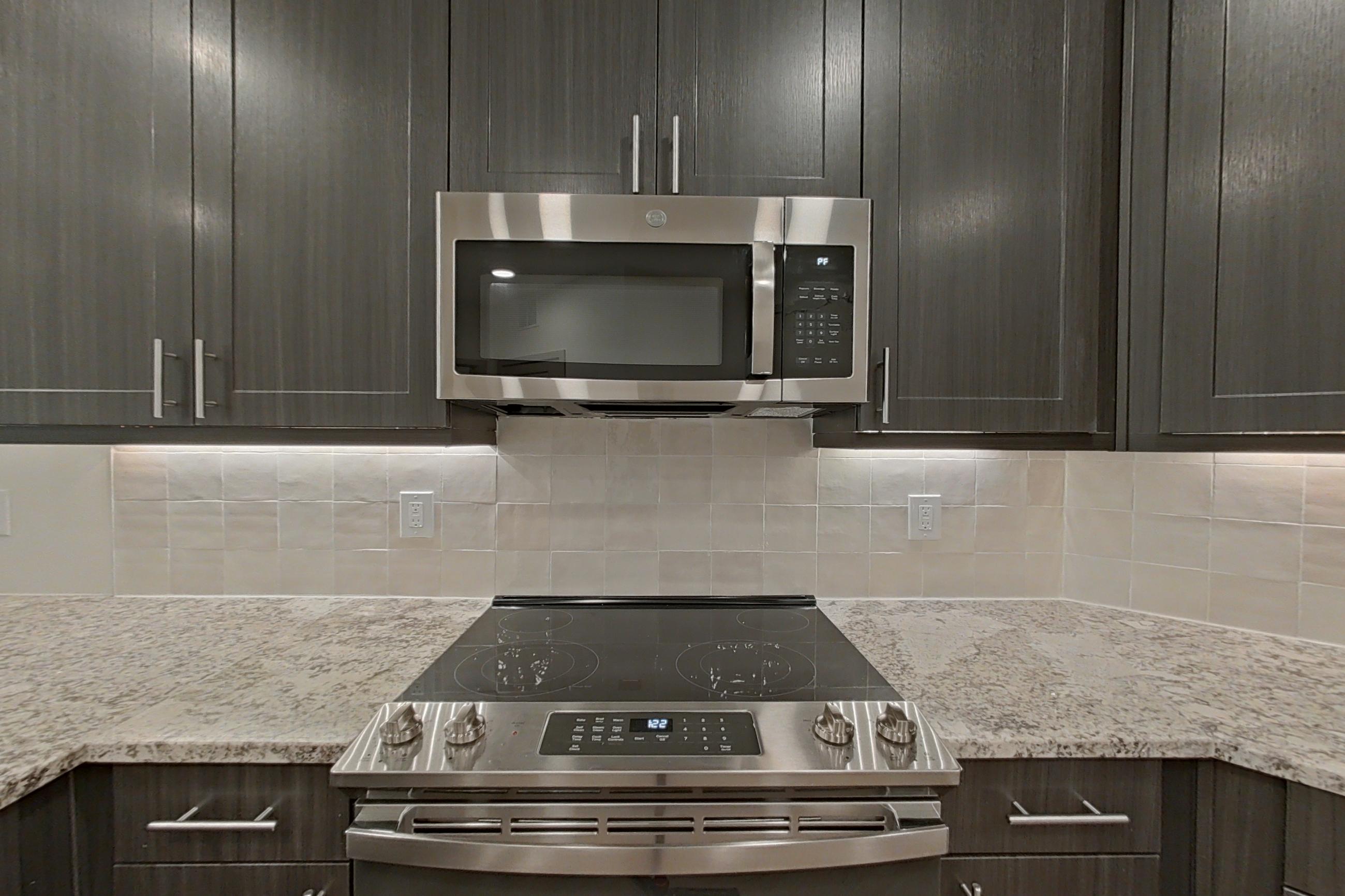 Kitchen with dishwasher, electric range, light stone countertops, light wood-finish flooring, microwave, oven/range, refrigerator, stainless steel appliances, and water & ice dispenser, Apartment 1302 at Merit Lewisville
