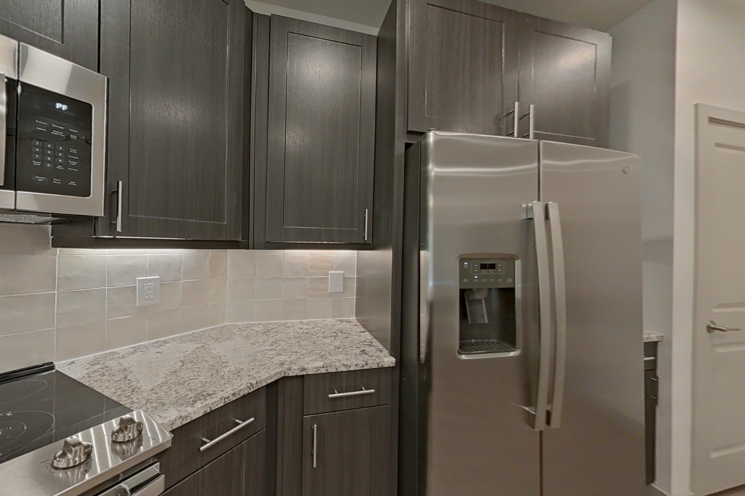 Kitchen with dishwasher, electric range, light stone countertops, light wood-finish flooring, microwave, oven/range, refrigerator, stainless steel appliances, and water & ice dispenser, Apartment 1302 at Merit Lewisville