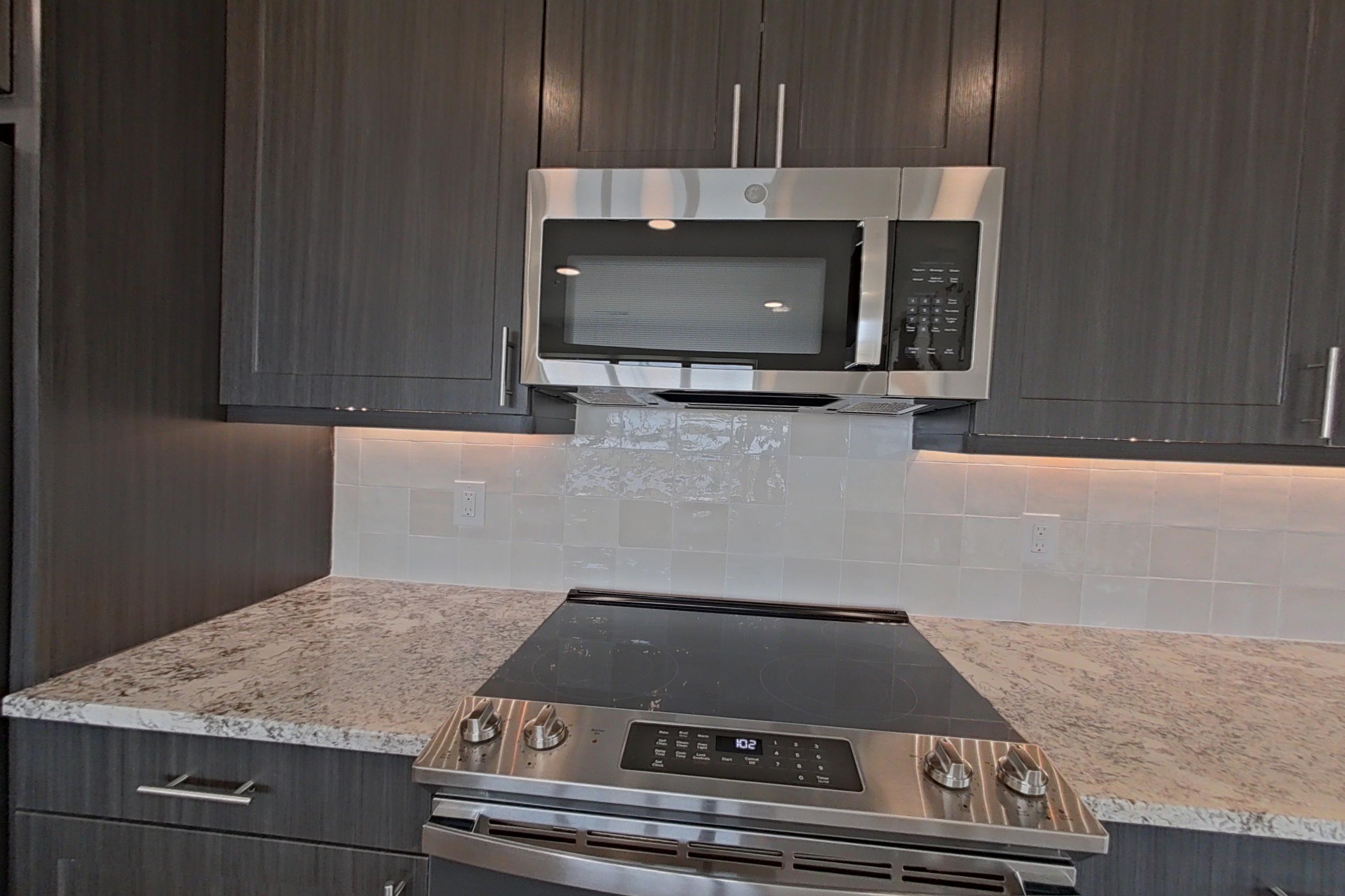 Kitchen with dishwasher, electric range, light stone countertops, light wood-finish flooring, microwave, oven/range, refrigerator, and stainless steel appliances, Apartment 1305 at Merit Lewisville