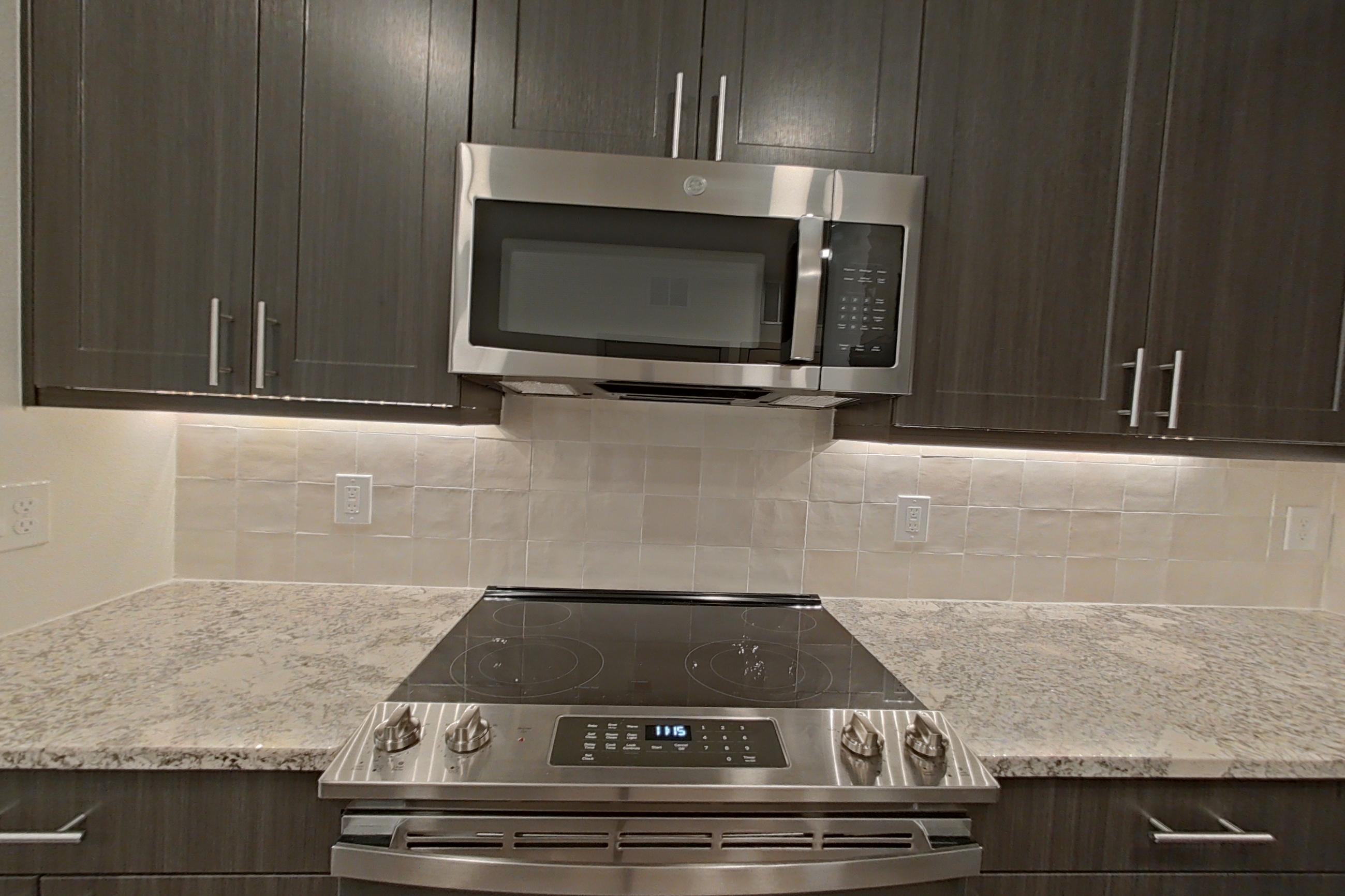 Kitchen with electric range, light stone countertops, light wood-finish flooring, microwave, oven/range, refrigerator, stainless steel appliances, and water & ice dispenser, Apartment 1310 at Merit Lewisville