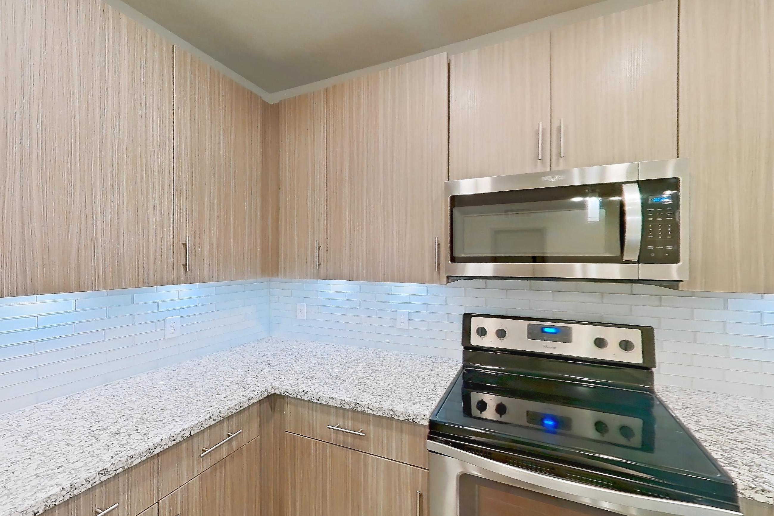 Kitchen with electric range, kitchen island, light countertops, light stone countertops, light wood-finish flooring, microwave, oven/range, refrigerator, stainless steel appliances, and water & ice dispenser, Apartment B3-102 at Lantower Grande Flats