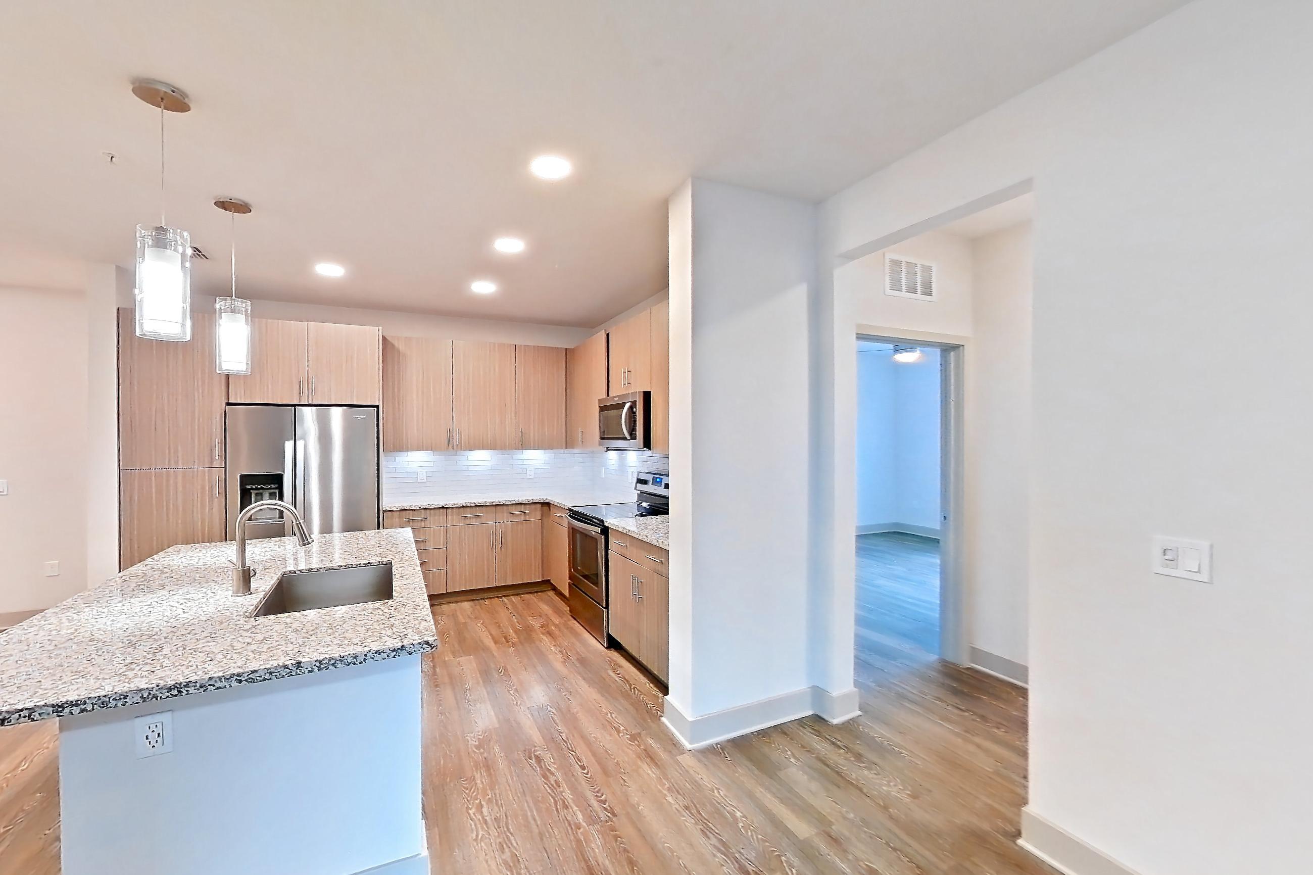 Living room with dark wood-finish flooring, electric range, kitchen island, light stone countertops, light wood-finish flooring, oven/range, refrigerator, stainless steel appliances, and water & ice dispenser, Apartment B3-102 at Lantower Grande Flats