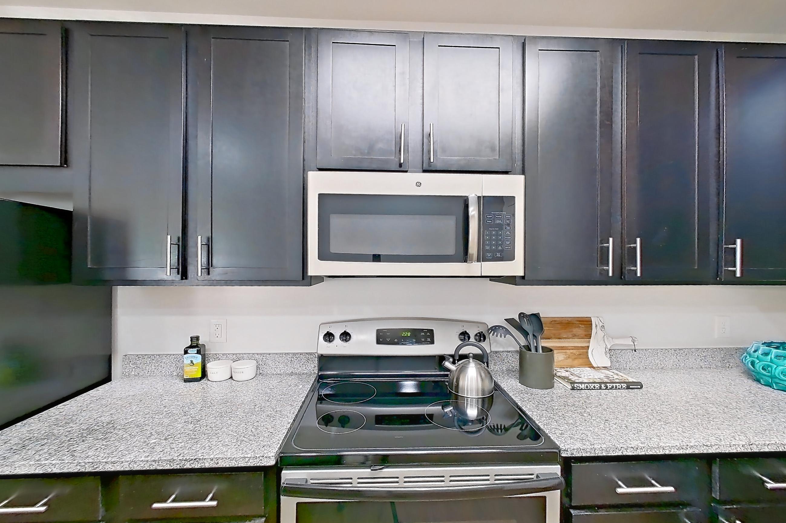 Kitchen with black appliances, dark wood-finish flooring, electric range, light countertops, light stone countertops, light wood-finish flooring, microwave, oven/range, refrigerator, and stainless steel appliances, Apartment 208 at Realm at Patterson Place