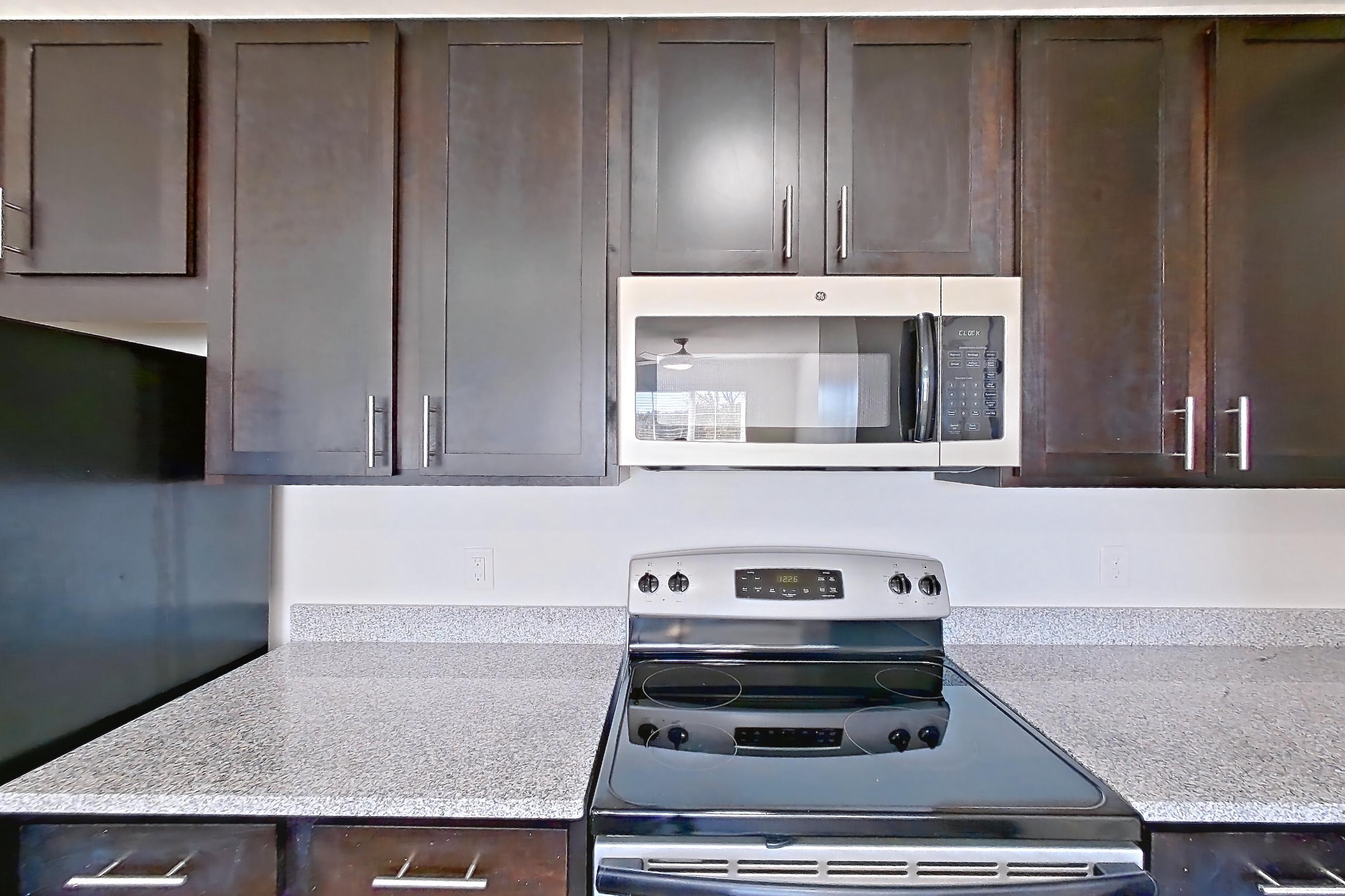 Kitchen with dark wood-finish flooring, dishwasher, electric range, light countertops, light stone countertops, light wood-finish flooring, microwave, oven/range, refrigerator, and stainless steel appliances, Apartment 101 at Realm at Patterson Place