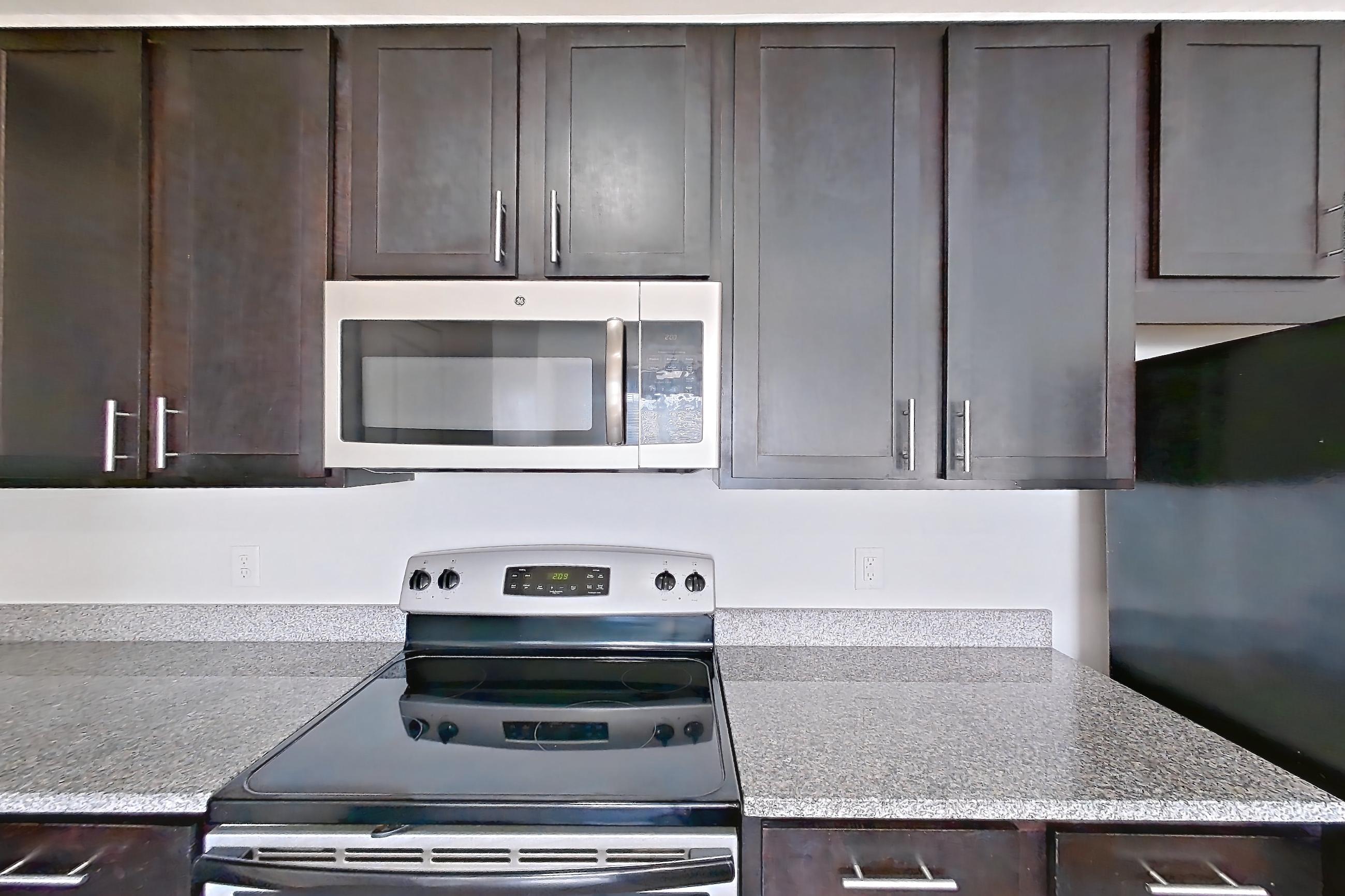Kitchen with dark wood-finish flooring, dishwasher, electric range, light countertops, light stone countertops, light wood-finish flooring, microwave, oven/range, refrigerator, and stainless steel appliances, Apartment 306 at Realm at Patterson Place
