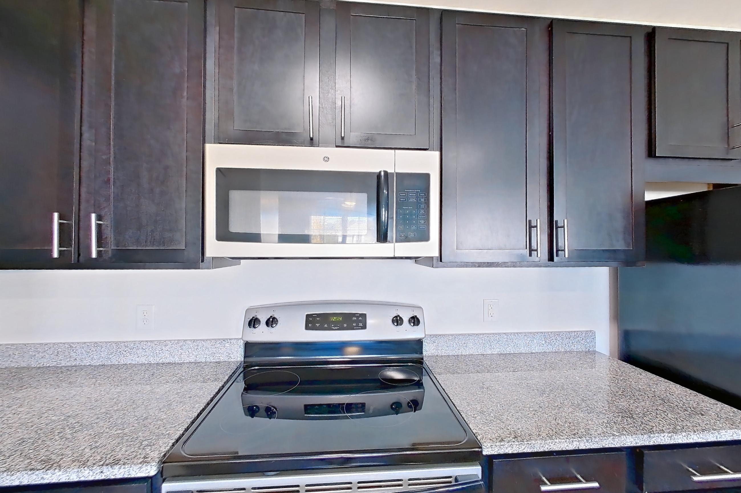 Kitchen with dark wood-finish flooring, dishwasher, electric range, kitchen island, light countertops, light stone countertops, light wood-finish flooring, microwave, oven/range, refrigerator, and stainless steel appliances, Apartment 303 at Realm at Patterson Place