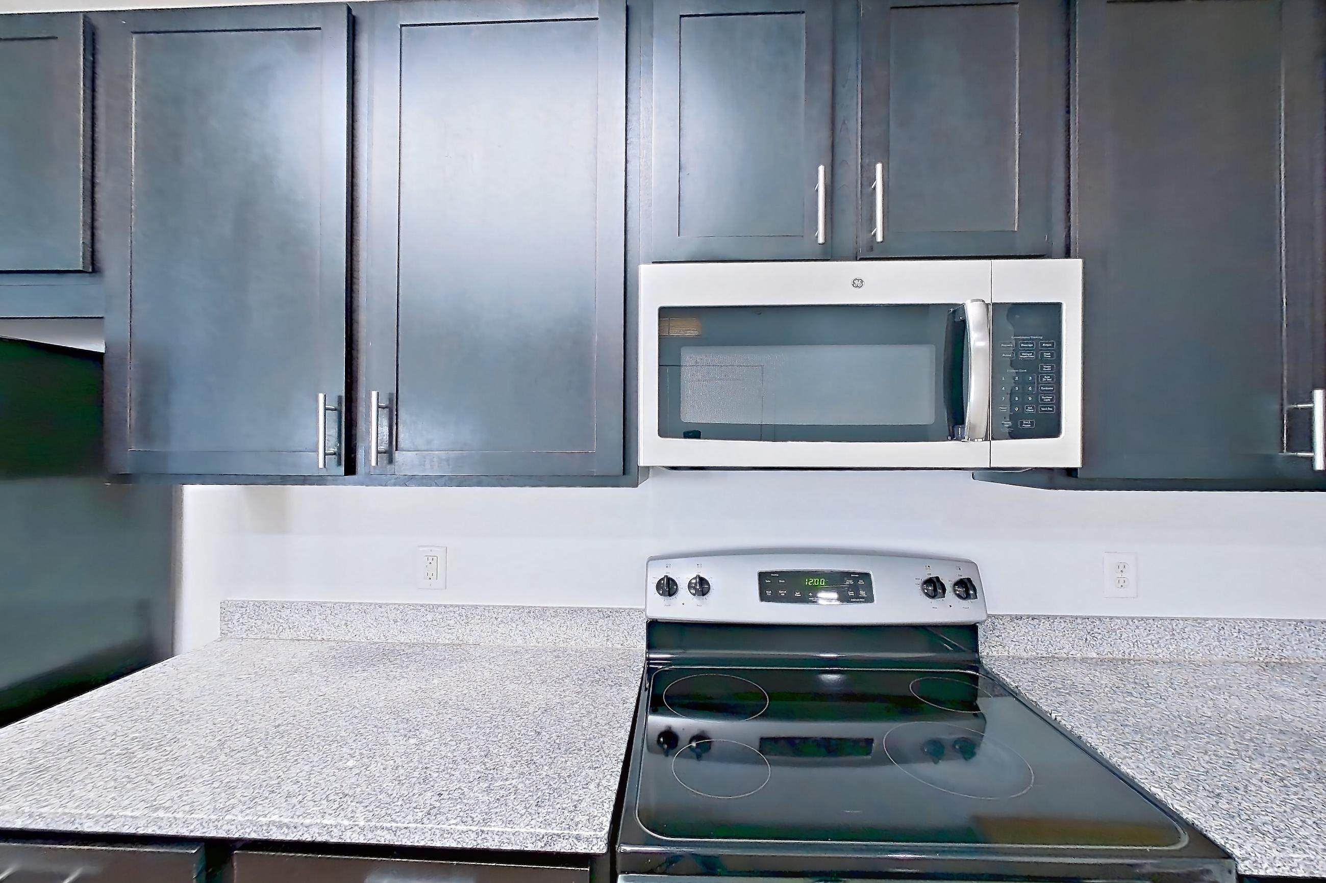 Kitchen with dark wood-finish flooring, dishwasher, electric range, light countertops, light stone countertops, light wood-finish flooring, microwave, oven/range, refrigerator, and stainless steel appliances, Apartment 208 at Realm at Patterson Place
