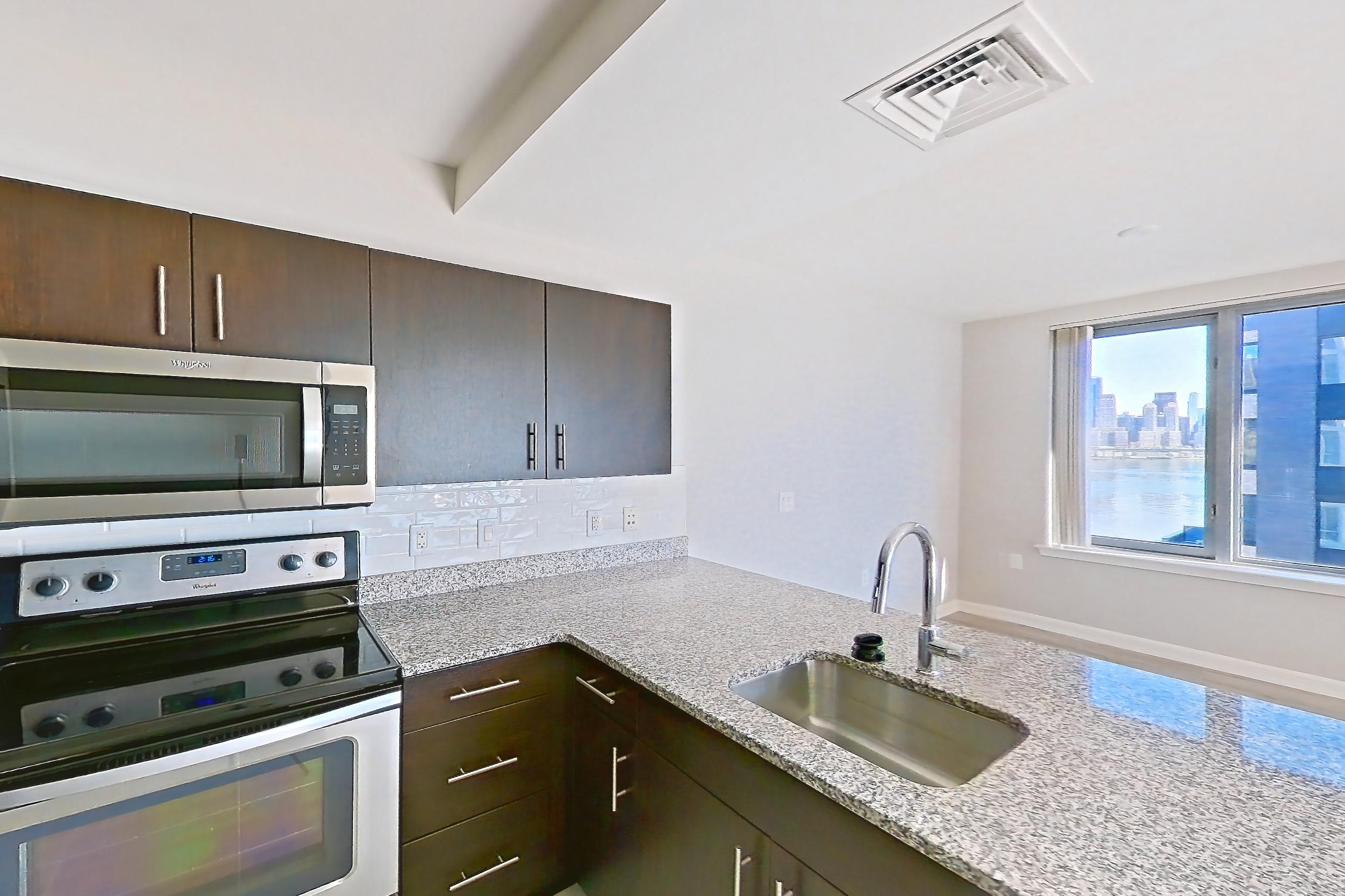 Kitchen with dark stone countertops, dishwasher, electric range, light stone countertops, light tile flooring, microwave, oven/range, refrigerator, and stainless steel appliances, Apartment RT-0922 at RiverTrace at Port Imperial