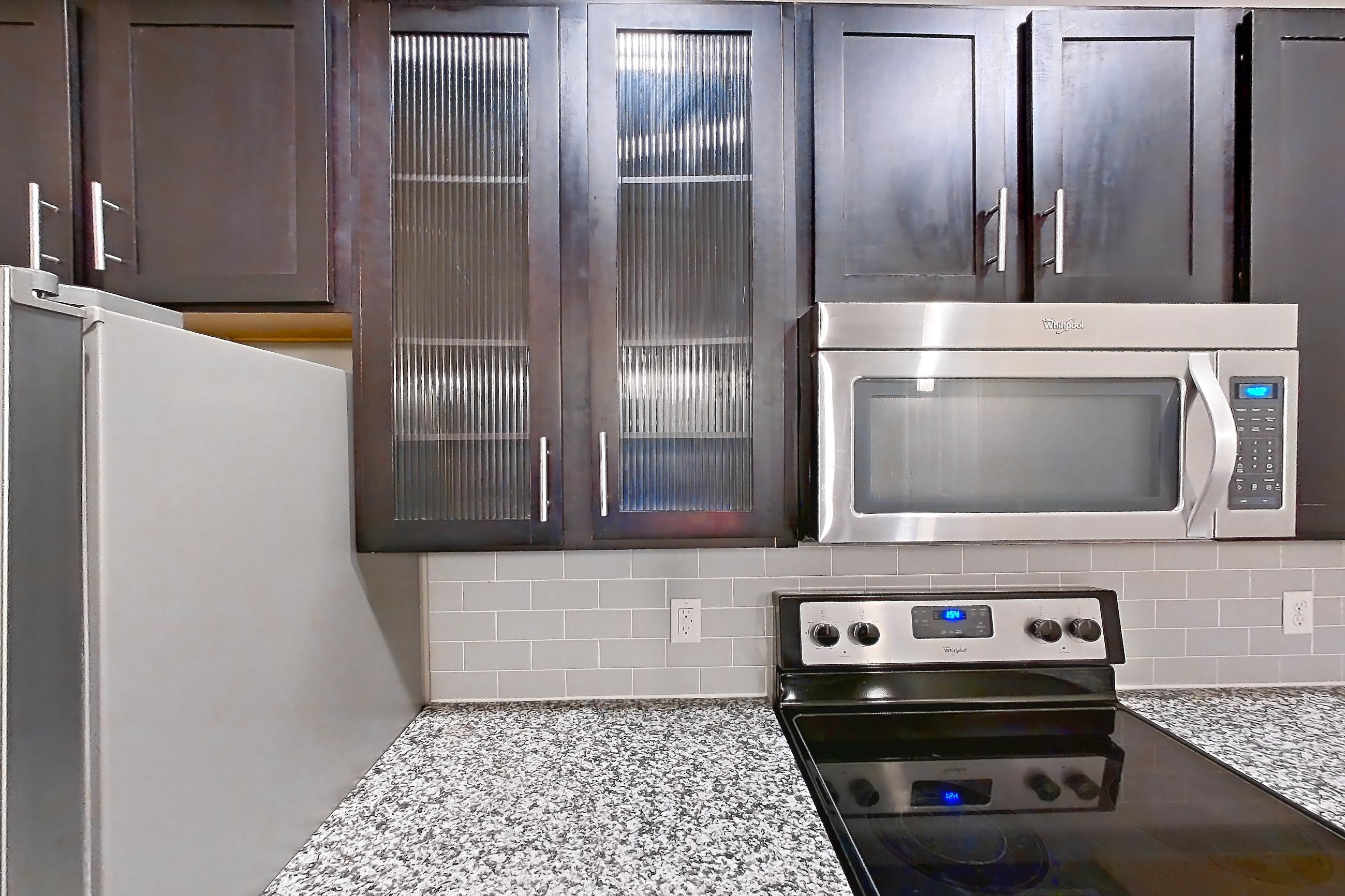 Kitchen with dark wood-finish flooring, dishwasher, electric range, light countertops, light stone countertops, light wood-finish flooring, microwave, oven/range, refrigerator, and stainless steel appliances, Apartment 0718 at Marquis at Barton Trails II