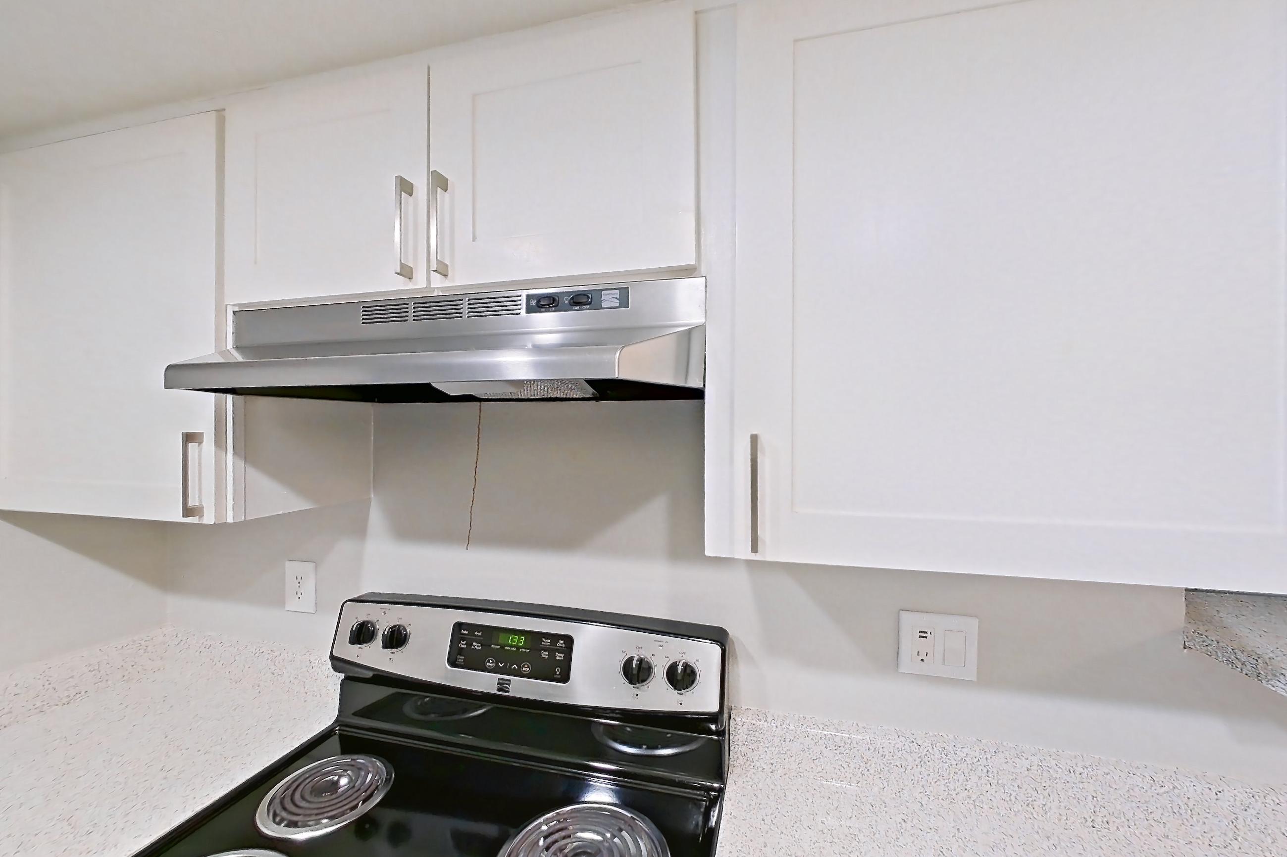 Kitchen with black appliances, dark wood-finish flooring, dishwasher, electric range, light countertops, light stone countertops, light wood-finish flooring, oven/range, range hood, refrigerator, and stainless steel appliances, Apartment 10 at Avana Rancho Cucamonga