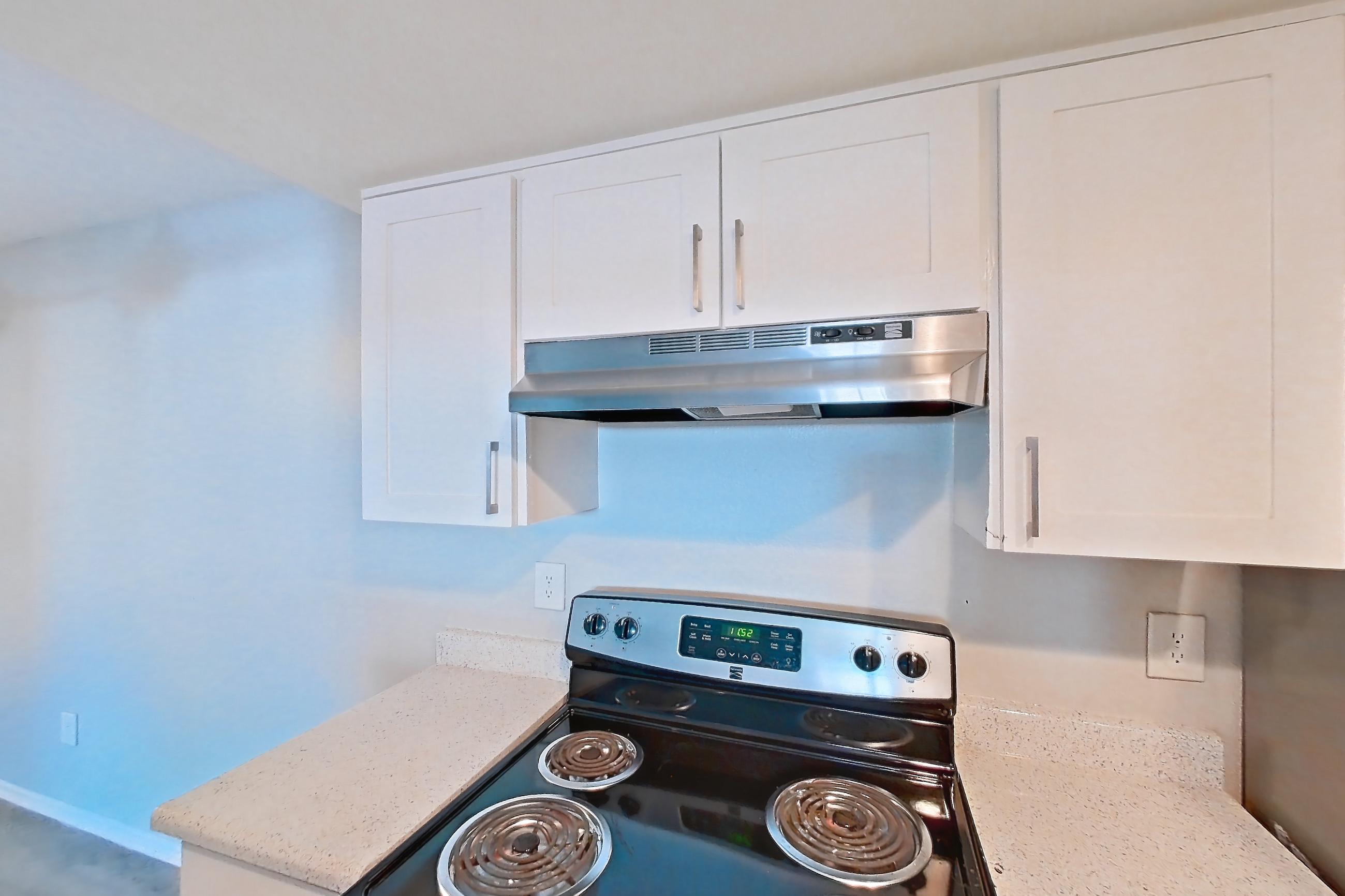 Kitchen with black appliances, electric range, light countertops, light stone countertops, light wood-finish flooring, oven/range, range hood, refrigerator, stainless steel appliances, and white appliances, Apartment 16 at Avana Rancho Cucamonga