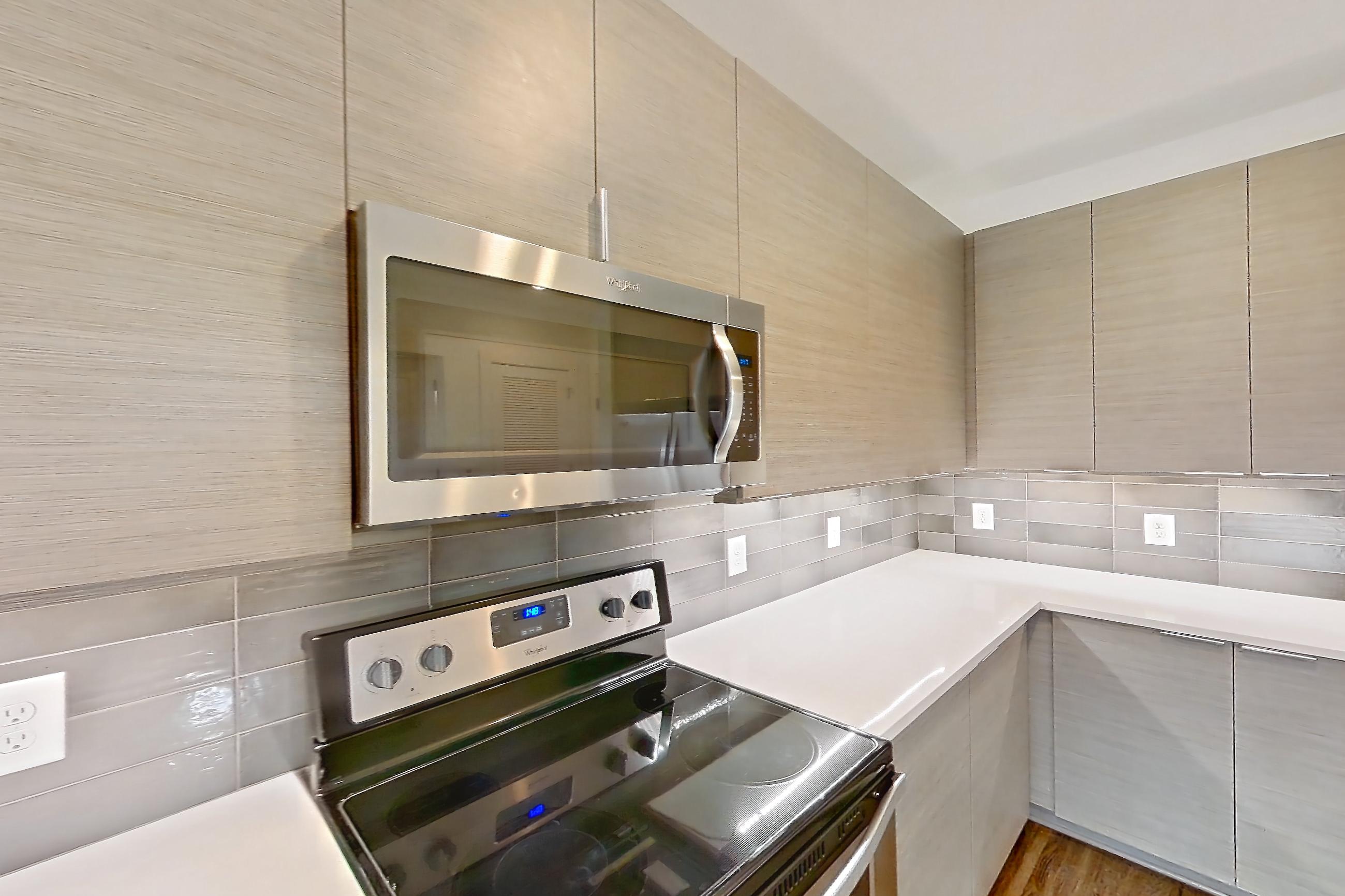 Kitchen with dark wood-finish flooring, dishwasher, electric range, kitchen island, light countertops, light stone countertops, microwave, oven/range, refrigerator, stainless steel appliances, and water & ice dispenser, Apartment 445 at Cortland at the Battery
