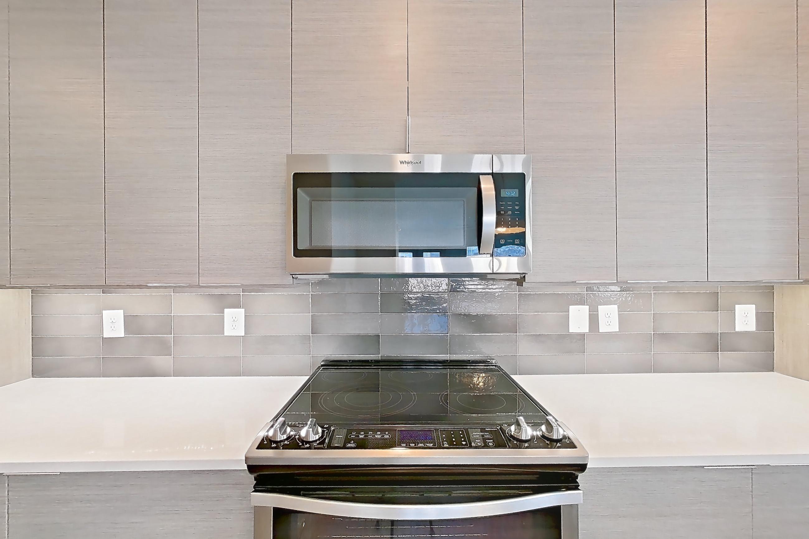 Kitchen with dark wood-finish flooring, electric range, kitchen island, light countertops, light stone countertops, microwave, oven/range, refrigerator, and stainless steel appliances, Apartment 432 at Cortland at the Battery