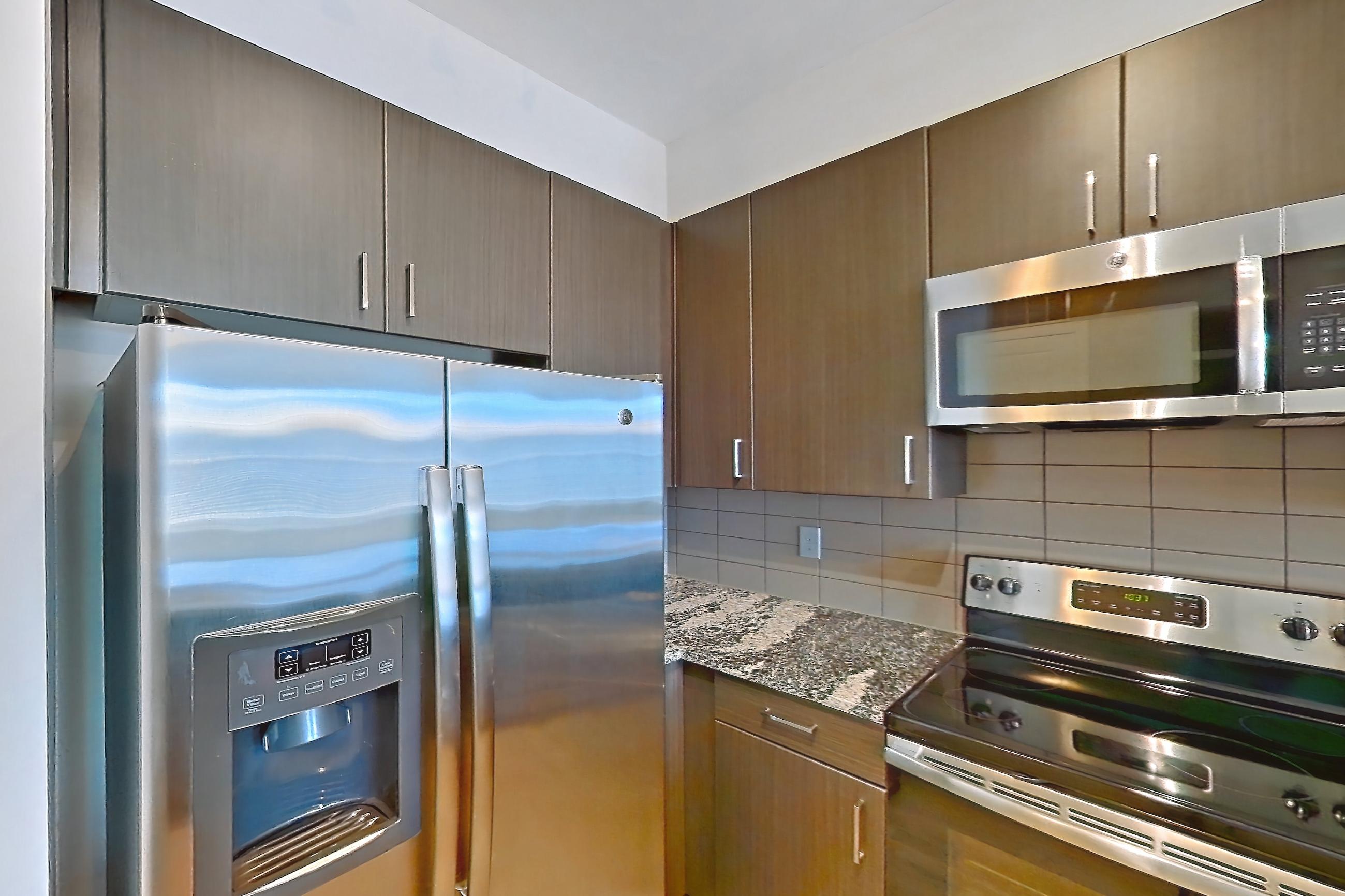 Kitchen with dark stone countertops, dishwasher, electric range, light stone countertops, light wood-finish flooring, microwave, oven/range, refrigerator, stainless steel appliances, and water & ice dispenser, Apartment 8305 at Cortland Wren Northlake