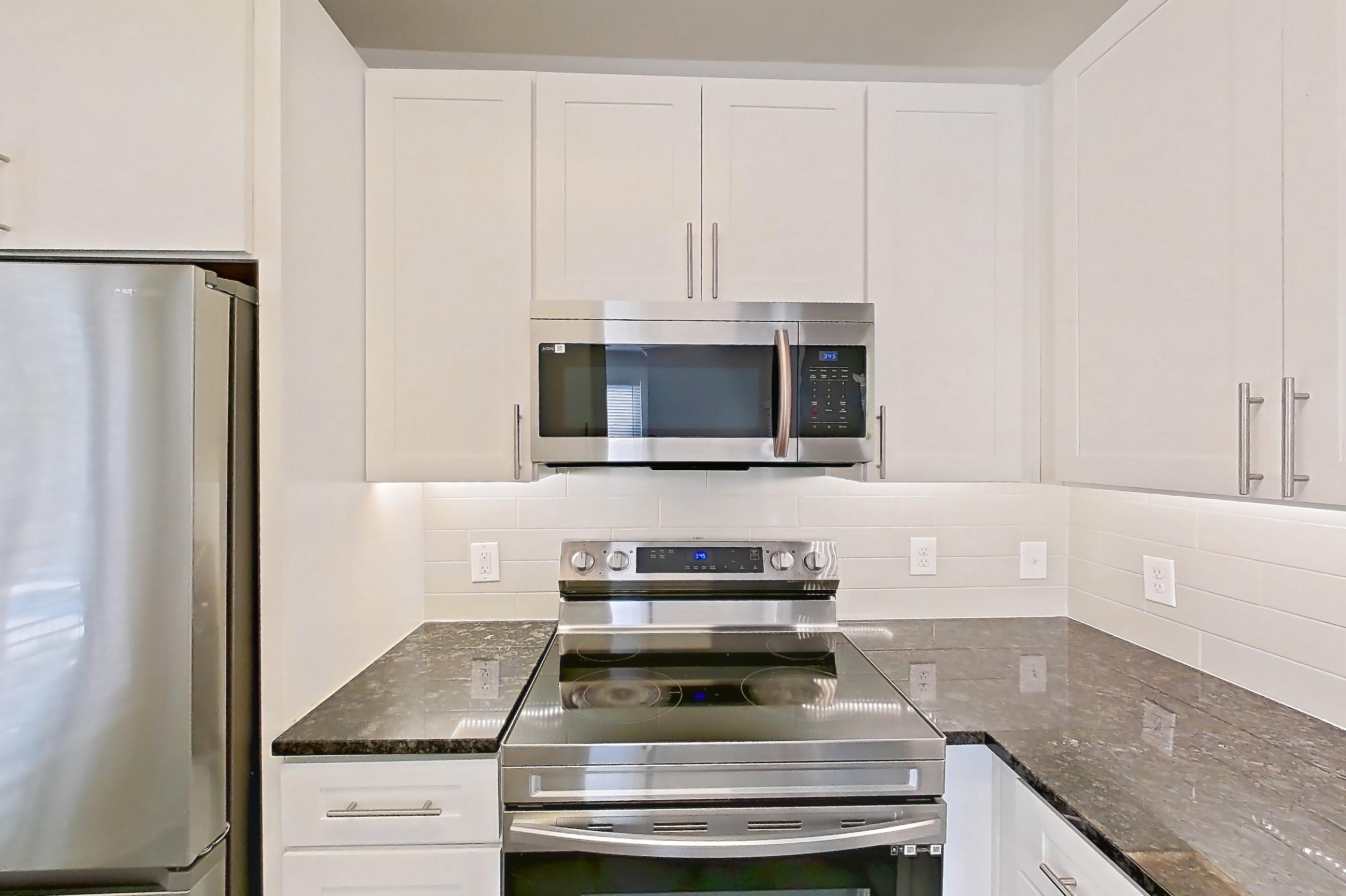 Kitchen with dark stone countertops, dark wood-finish flooring, dishwasher, electric range, light countertops, microwave, oven/range, refrigerator, and stainless steel appliances, Apartment 02432 at Cortland West Plano