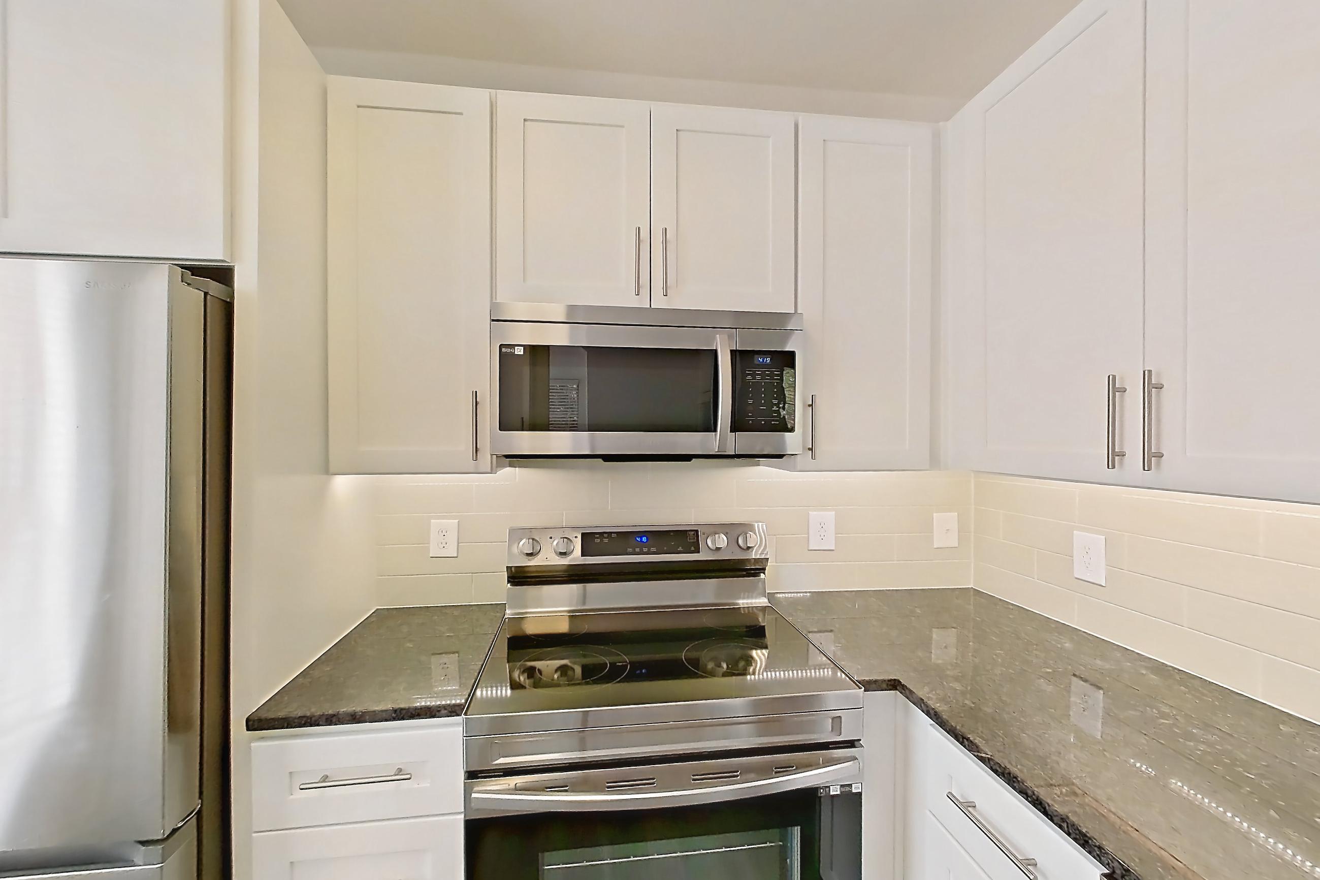 Kitchen with dark stone countertops, dark wood-finish flooring, dishwasher, electric range, light countertops, microwave, oven/range, refrigerator, stainless steel appliances, and white appliances, Apartment 03025 at Cortland West Plano