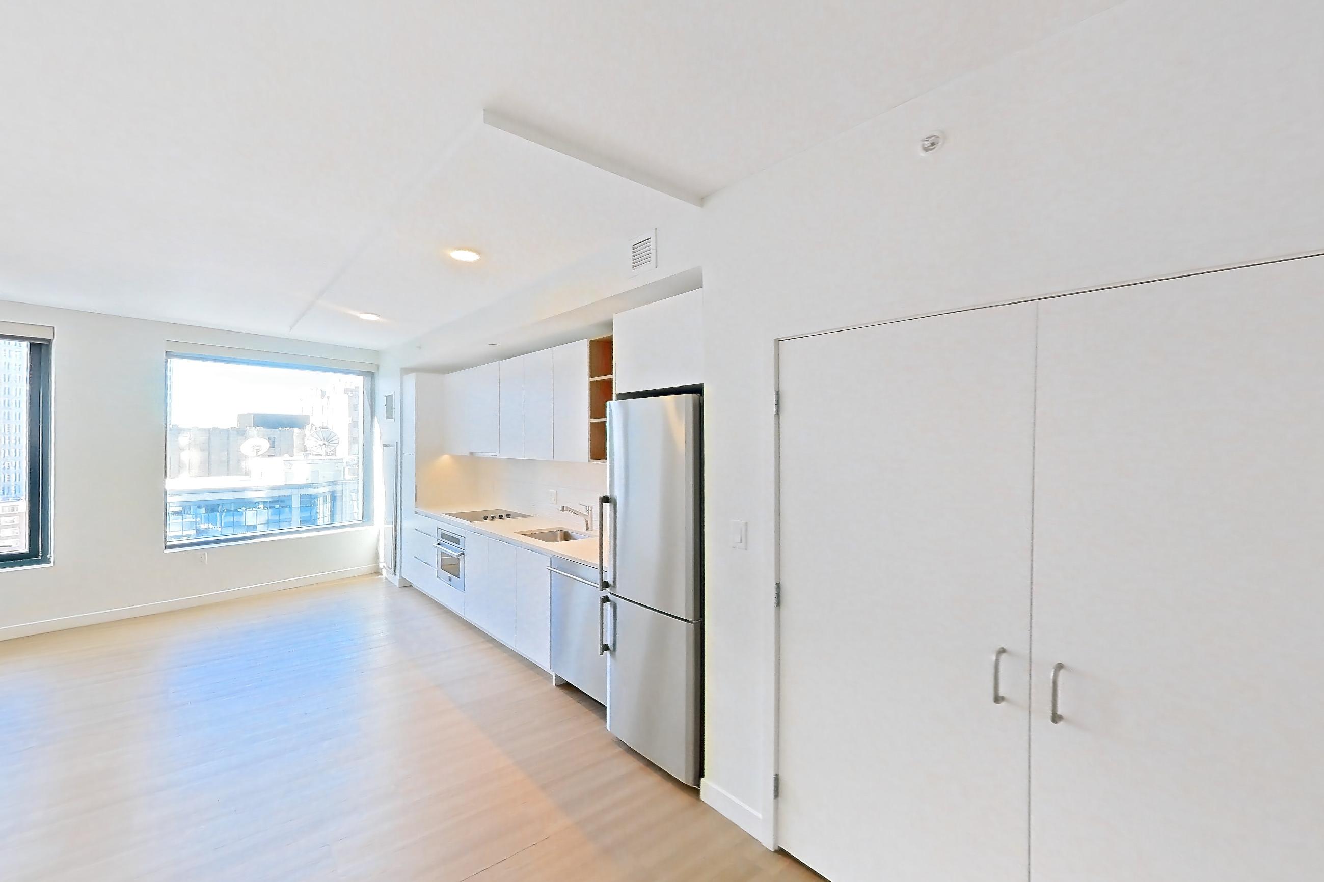 Living room with light wood-finish flooring, refrigerator, and stainless steel appliances, Apartment 1112 at The Sudbury