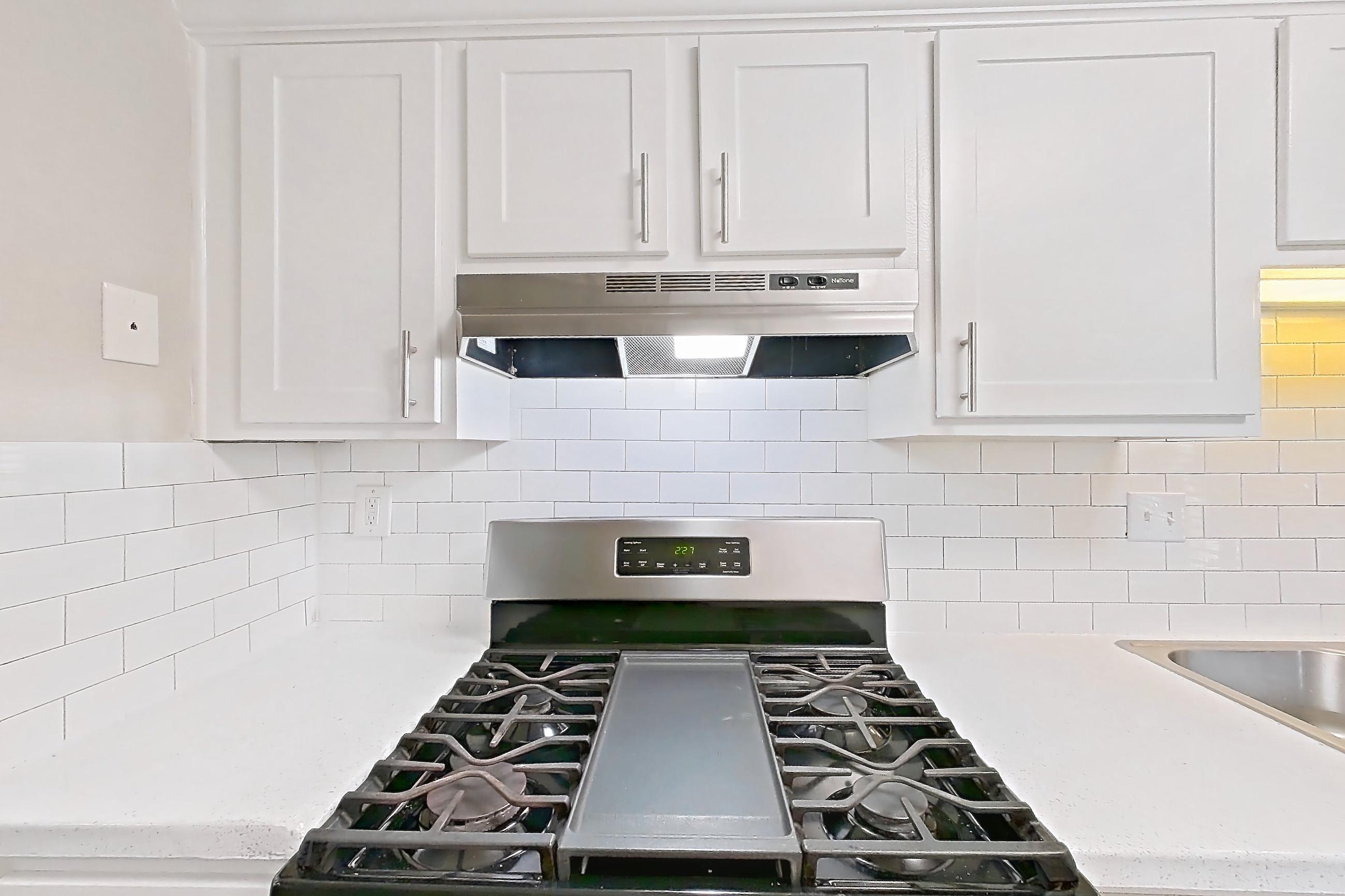 Kitchen with dishwasher, gas range, light countertops, light stone countertops, light wood-finish flooring, oven/range, range hood, refrigerator, and stainless steel appliances, Apartment H1456T at The Legacy at Druid Hills