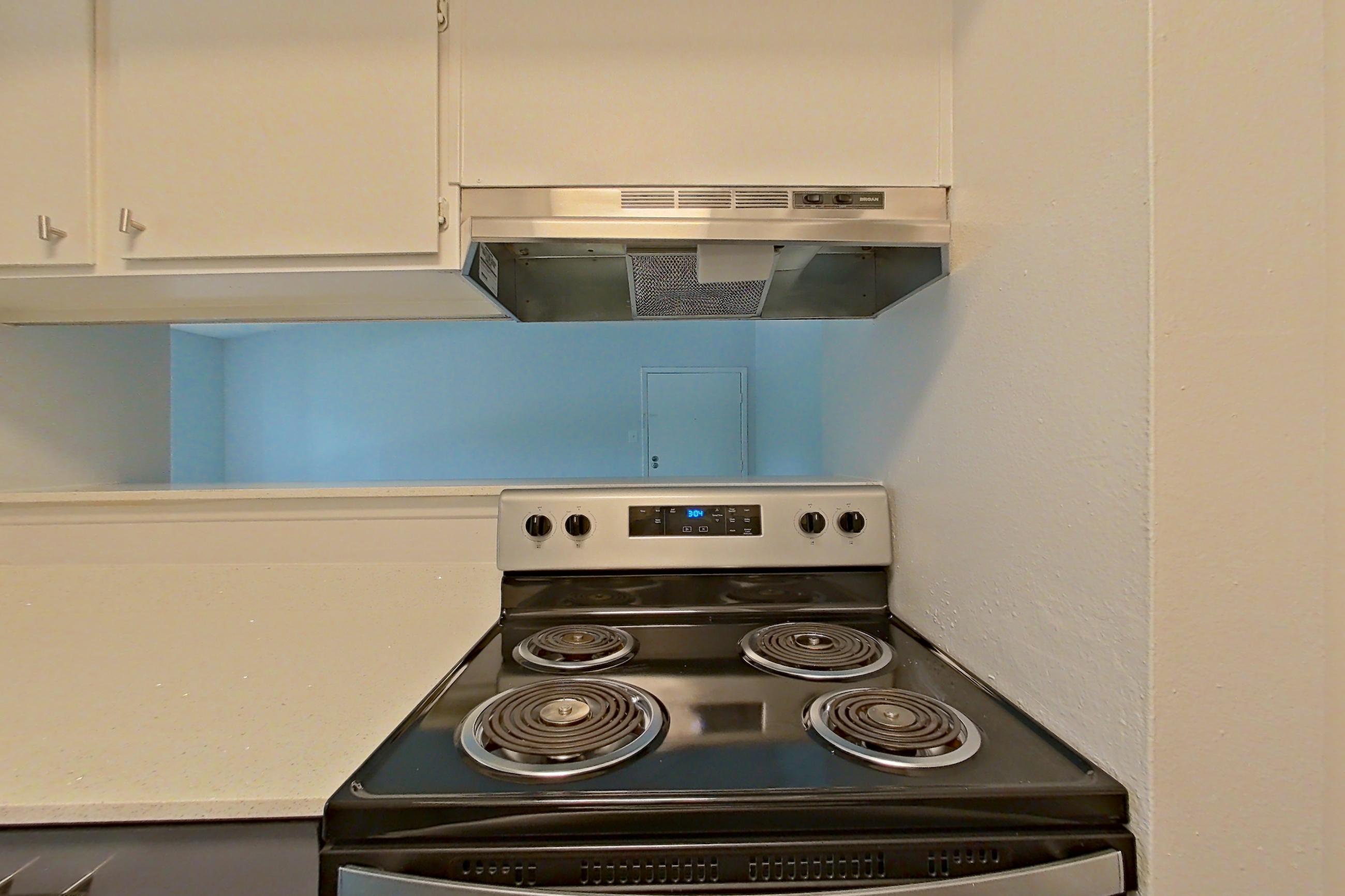 Kitchen with dark wood-finish flooring, dishwasher, electric range, light countertops, light stone countertops, oven/range, range hood, refrigerator, stainless steel appliances, and white appliances, Apartment 1904 at The Cove Apartments
