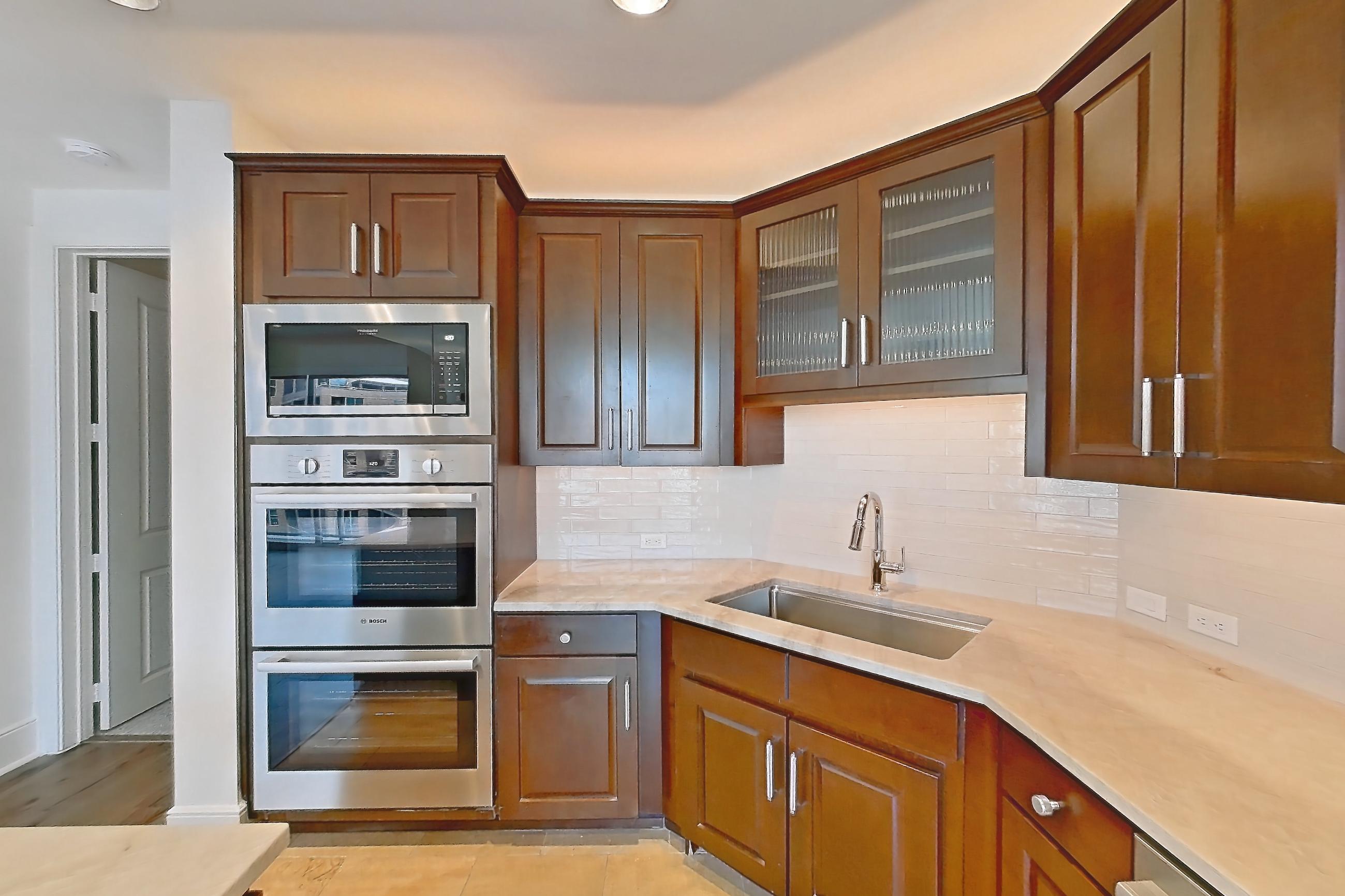 Kitchen with black appliances, built-in microwave, cooktop, dishwasher, double oven, electric cooktop, kitchen island, light countertops, light stone countertops, light tile flooring, light wood-finish flooring, range hood, refrigerator, stainless steel appliances, and water & ice dispenser, Apartment 1507 at Cortland on McKinney