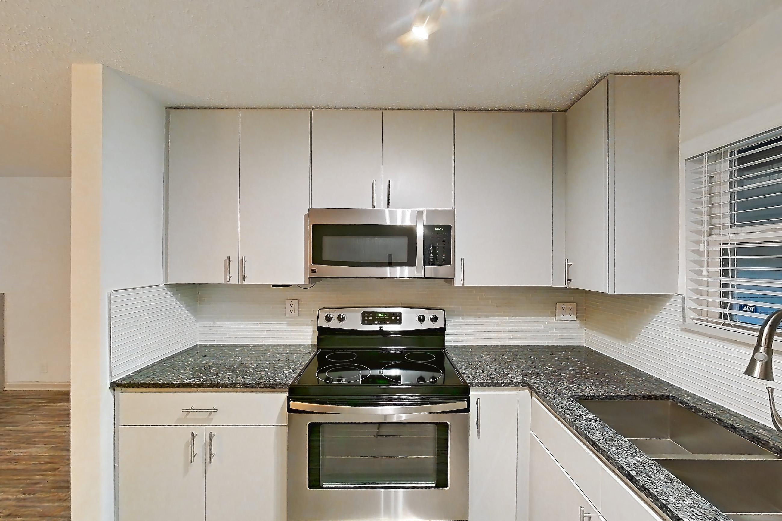 Kitchen with dark stone countertops, dishwasher, electric range, fireplace, light countertops, light wood-finish flooring, microwave, oven/range, refrigerator, and stainless steel appliances, Apartment 3 at Cortland Cotswold
