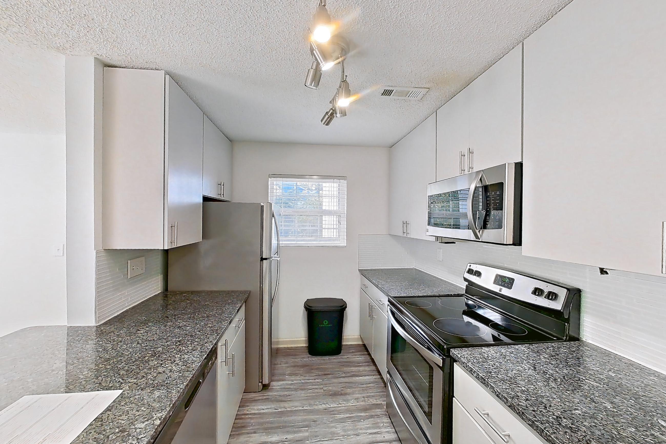 Kitchen with dark stone countertops, dishwasher, electric range, light carpet, light countertops, light wood-finish flooring, oven/range, refrigerator, and stainless steel appliances, Apartment 8 at Cortland Cotswold