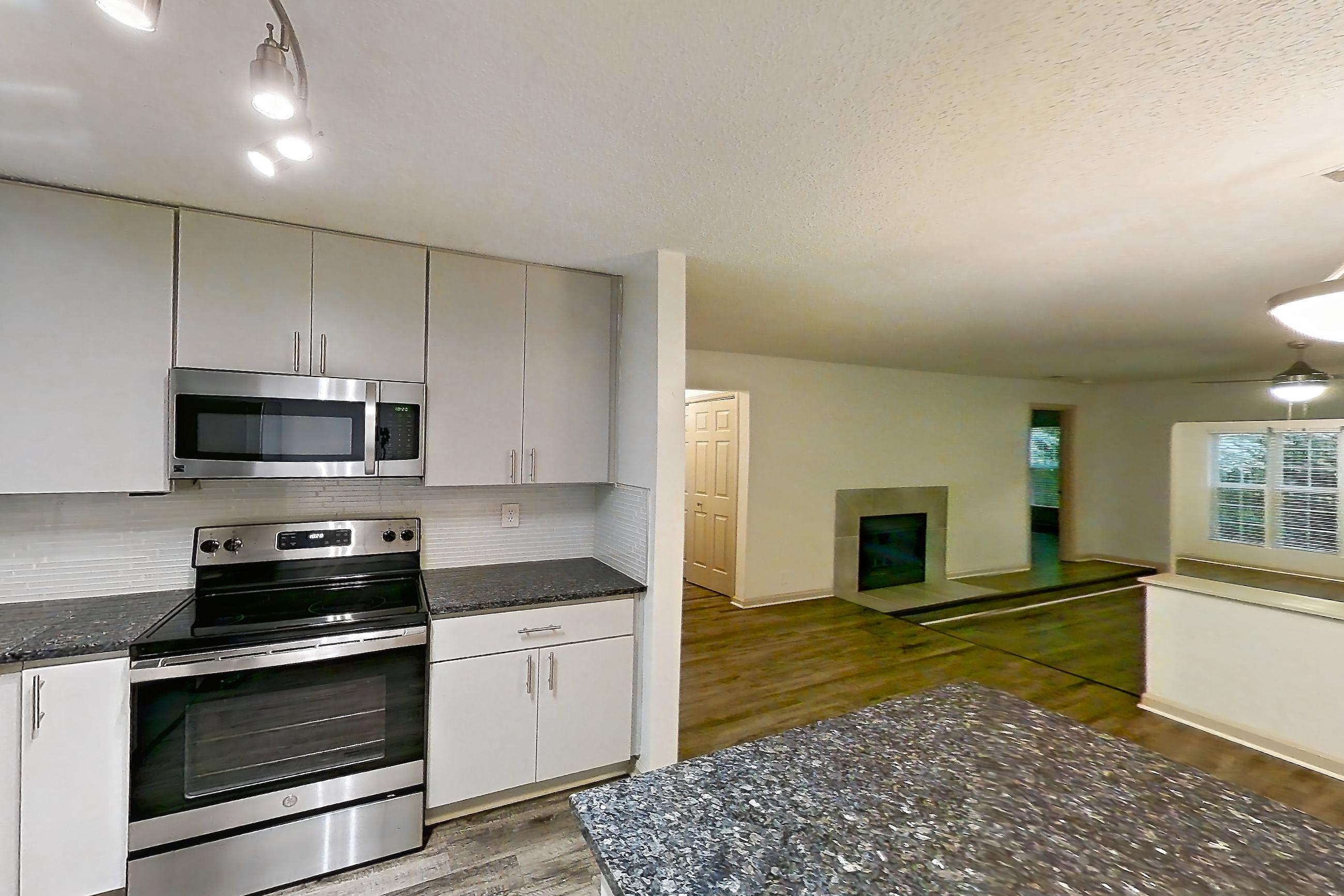 Kitchen with dark stone countertops, dishwasher, electric range, fireplace, light countertops, light wood-finish flooring, microwave, oven/range, refrigerator, stainless steel appliances, and tile fireplace, Apartment 2 at Cortland Cotswold