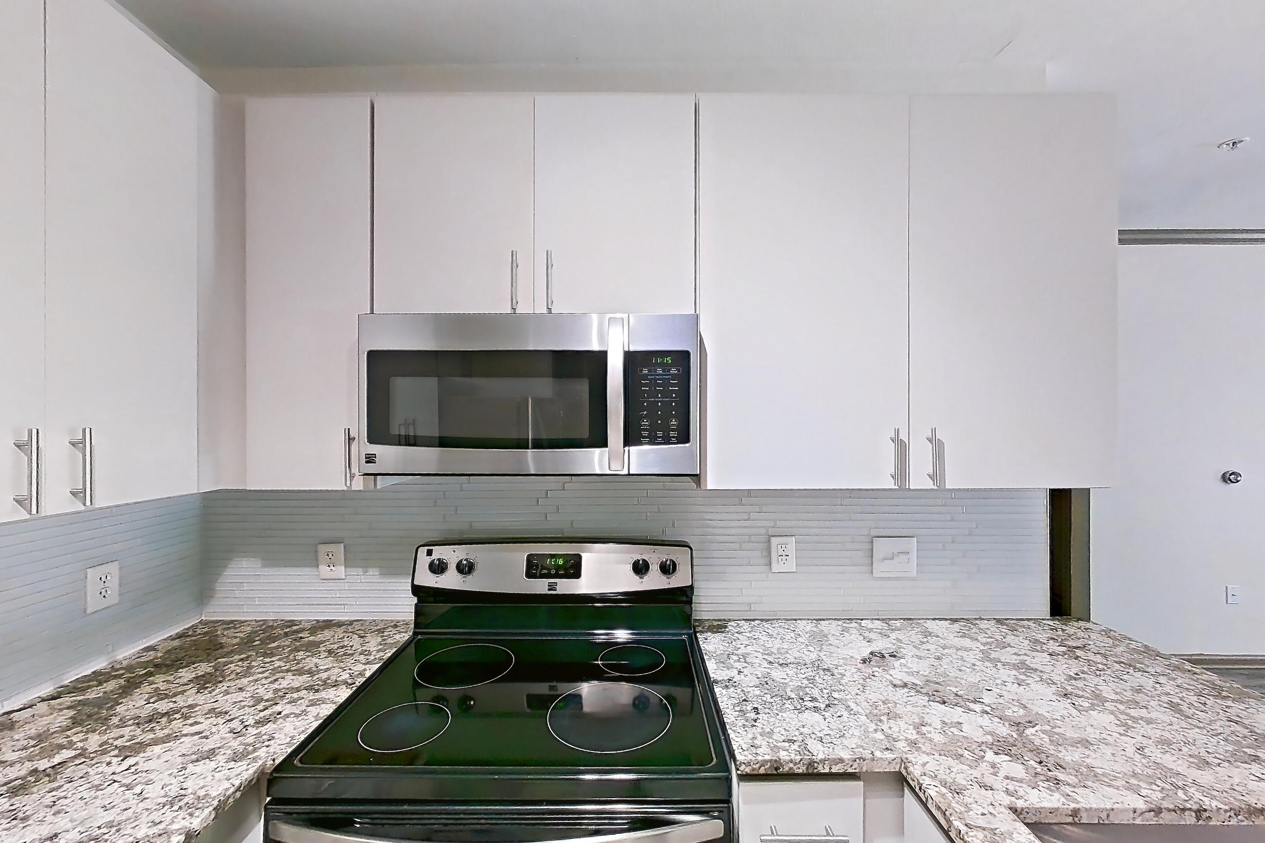 Kitchen with dishwasher, electric range, kitchen island, light countertops, light stone countertops, light wood-finish flooring, microwave, oven/range, refrigerator, and stainless steel appliances, Apartment 1317 at Cortland Seventy Seven