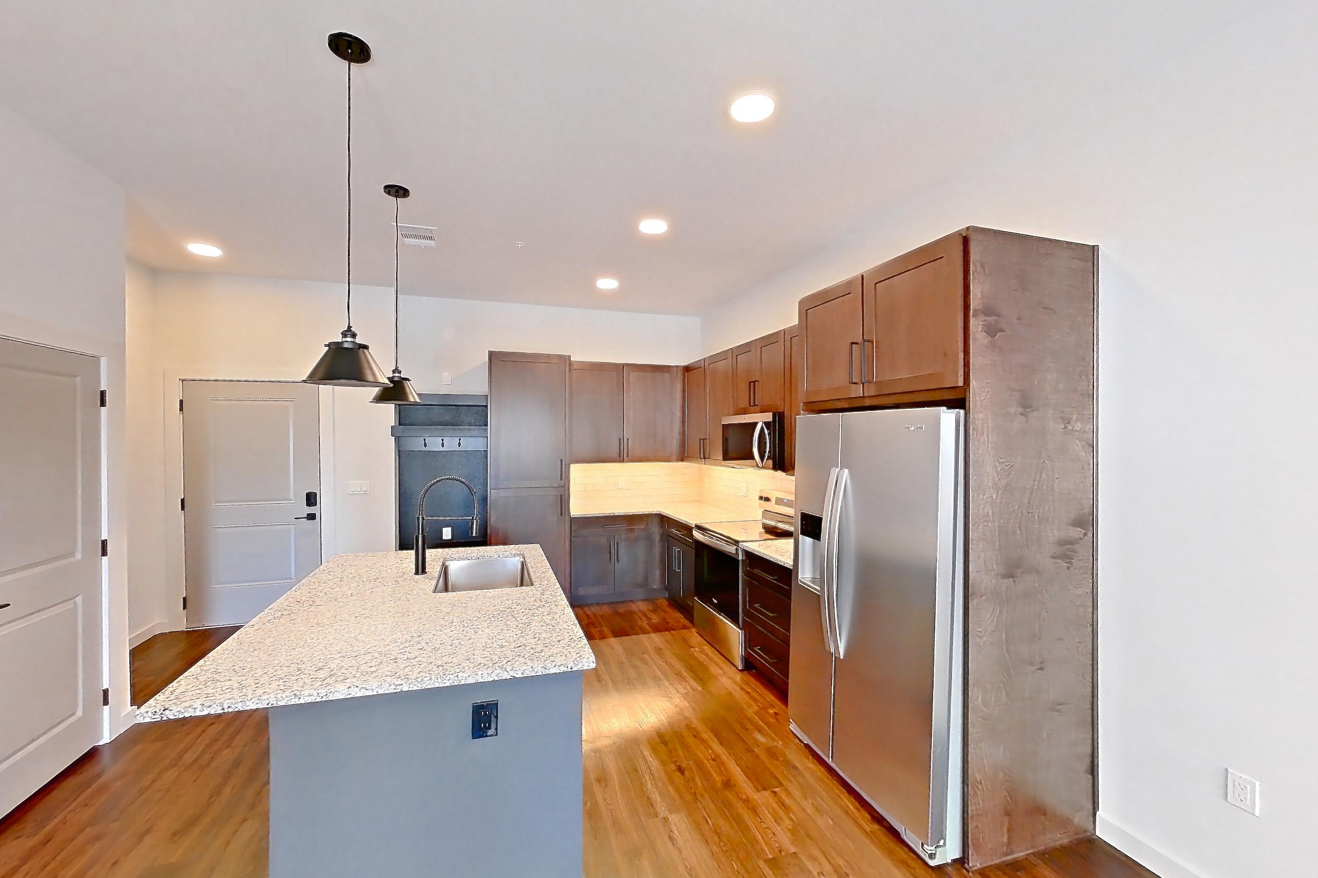 Living space with dark wood-finish flooring, electric range, kitchen island, light countertops, light stone countertops, oven/range, refrigerator, stainless steel appliances, and water & ice dispenser, Apartment 1-121 at Cortland Peterson