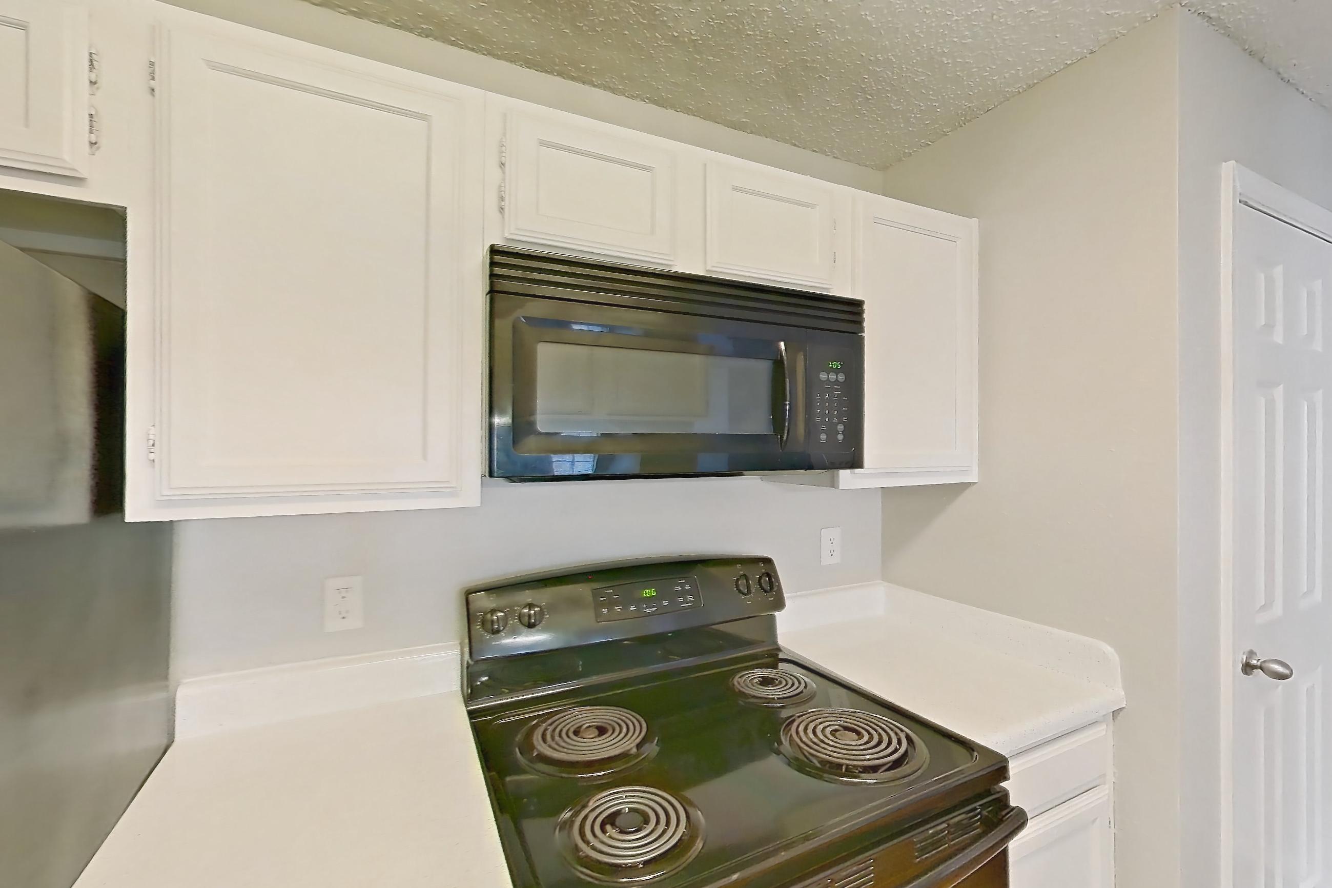 Kitchen with black appliances, dishwasher, electric range, fireplace, light countertops, light wood-finish flooring, microwave, oven/range, range hood, refrigerator, stainless steel appliances, and wood-finish flooring, Apartment 08-0821 at The Arbors of Las Colinas