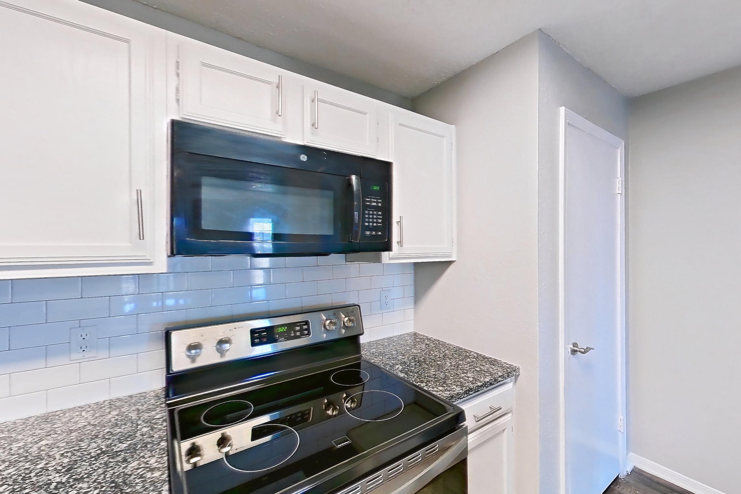 Kitchen with black appliances, dark stone countertops, dishwasher, electric range, light countertops, light wood-finish flooring, microwave, oven/range, refrigerator, and stainless steel appliances, Apartment 05-0521 at The Arbors of Las Colinas