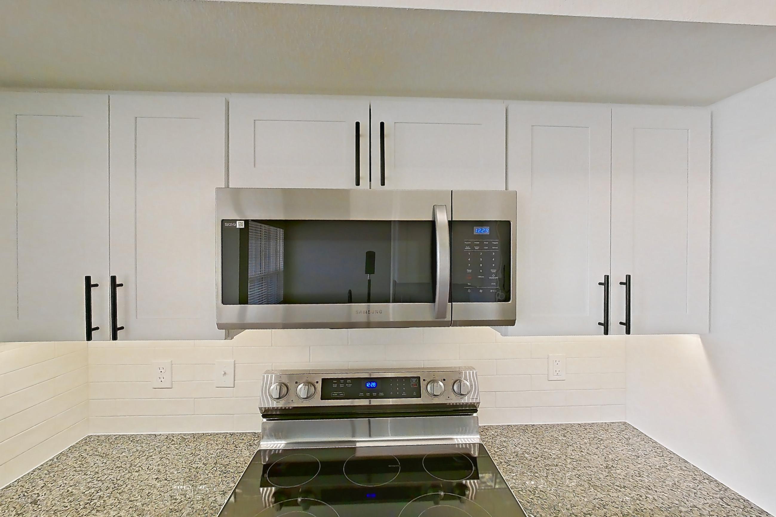 Kitchen with dark stone countertops, dishwasher, electric range, light countertops, light stone countertops, light wood-finish flooring, microwave, oven/range, refrigerator, and stainless steel appliances, Apartment 16-303 at Cortland Westminster