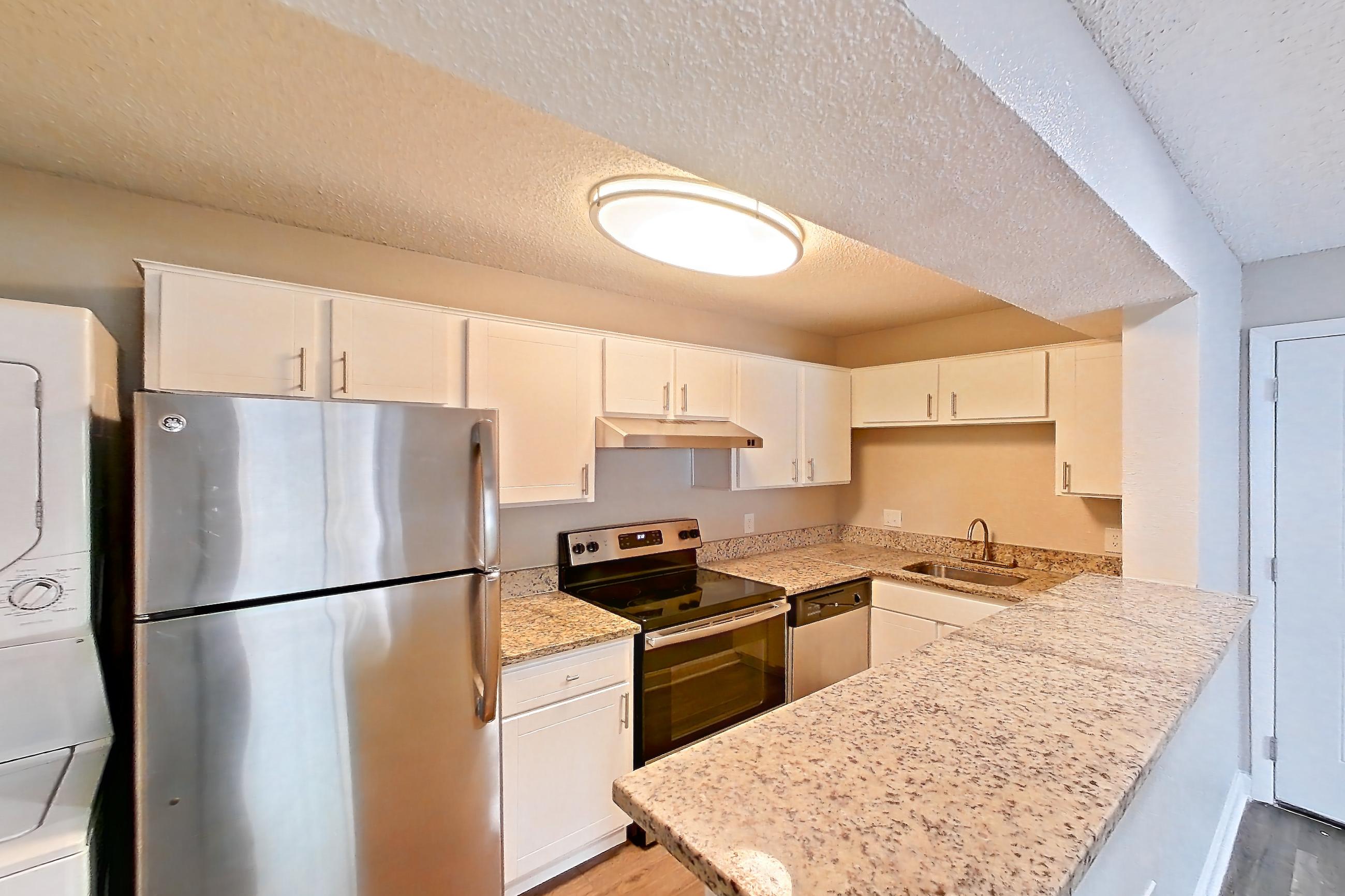 Kitchen entryway with dark wood-finish flooring, dishwasher, electric range, in-unit laundry, light countertops, light stone countertops, light wood-finish flooring, oven/range, refrigerator, and stainless steel appliances, Apartment 2103 at Cortland at the Hammocks