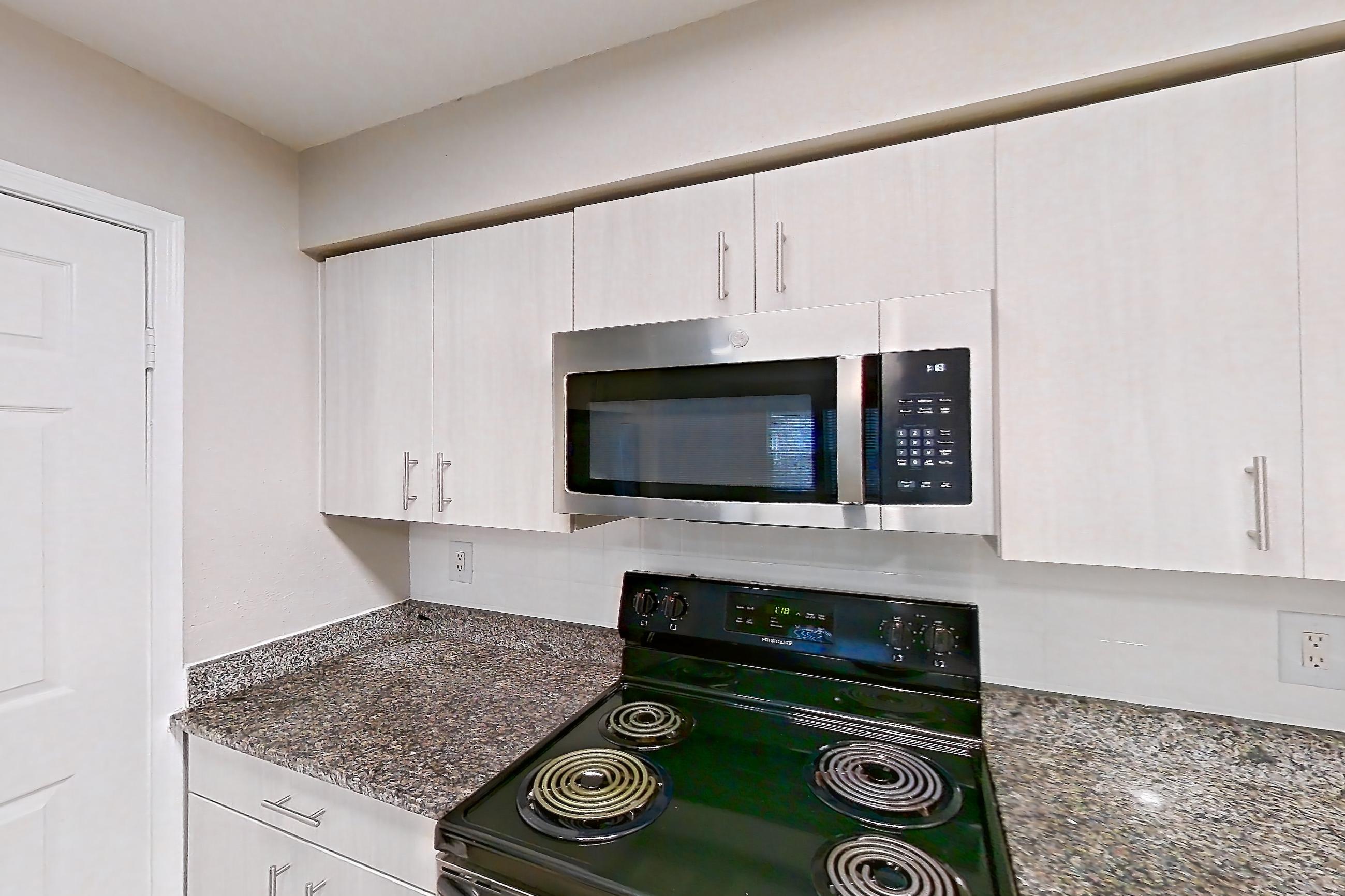 Kitchen with dark stone countertops, dishwasher, electric range, fireplace, light countertops, light stone countertops, light wood-finish flooring, microwave, oven/range, refrigerator, stainless steel appliances, and tile fireplace, Apartment 1101 at Cortland Van Dorn