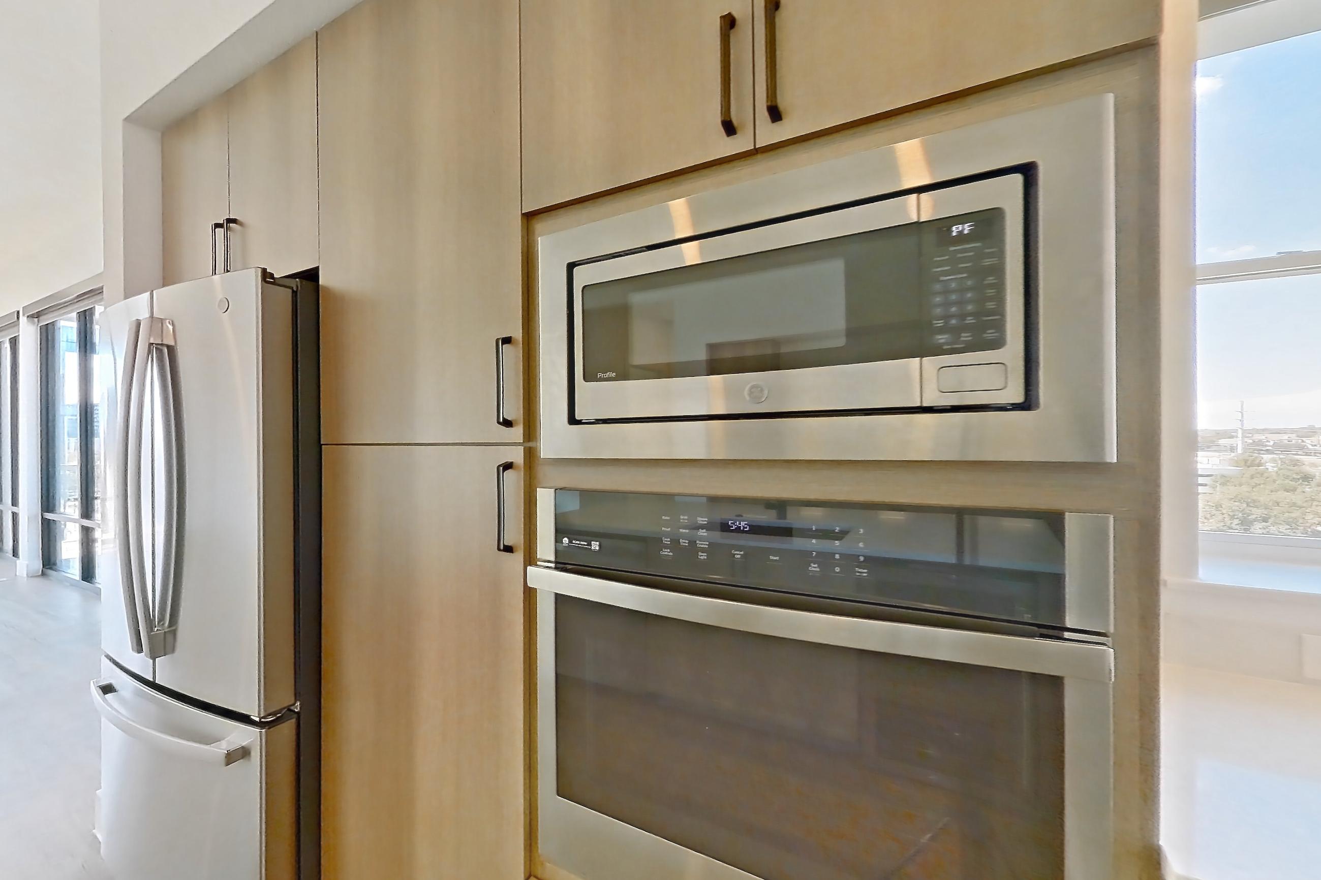 Kitchen with built-in microwave, dishwasher, light countertops, light stone countertops, light wood-finish flooring, microwave, oven/range, refrigerator, and stainless steel appliances, Apartment 703 at The Watson
