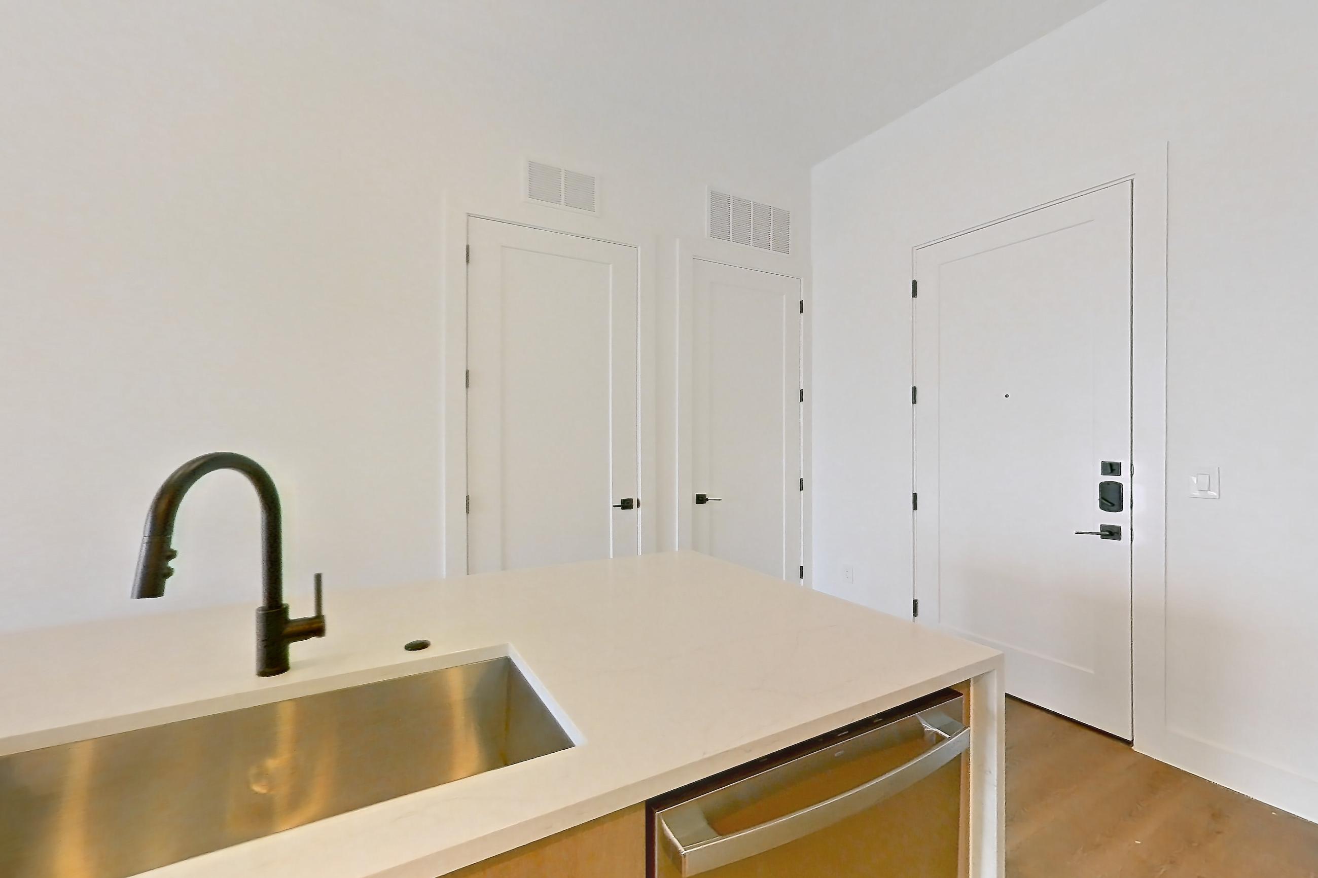 Kitchen with black appliances, cooktop, dishwasher, electric cooktop, kitchen island, light countertops, light stone countertops, light wood-finish flooring, range hood, refrigerator, and stainless steel appliances, Apartment 715 at The Watson
