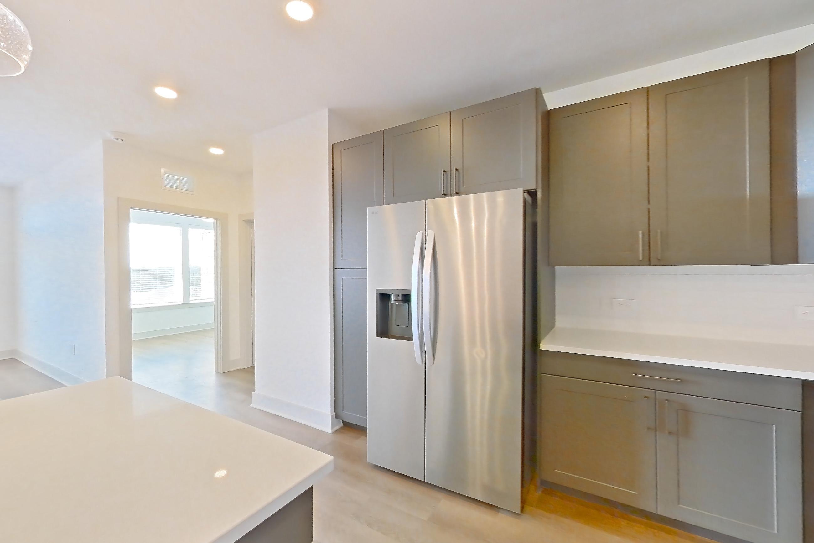 Kitchen with dishwasher, kitchen island, light countertops, light stone countertops, light wood-finish flooring, oven/range, refrigerator, stainless steel appliances, and water & ice dispenser, Apartment 2402 at Groves South Congress
