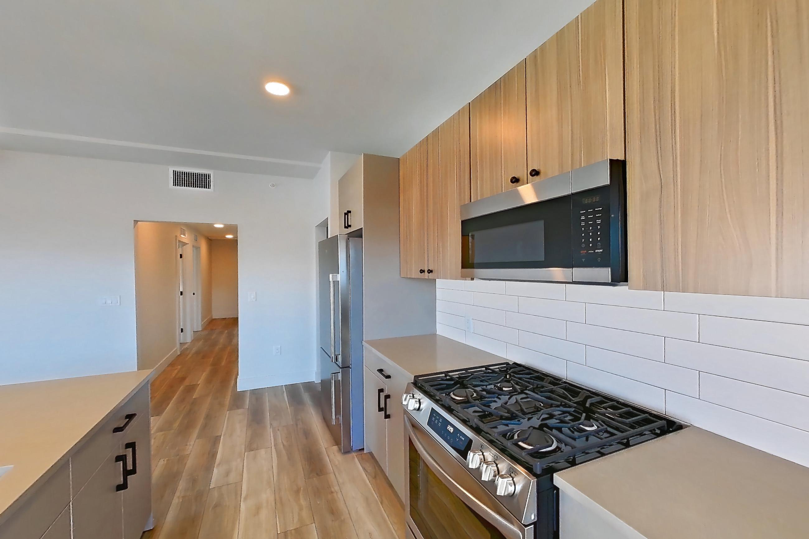 Kitchen with dishwasher, gas range, kitchen island, light countertops, light stone countertops, light wood-finish flooring, microwave, oven/range, refrigerator, and stainless steel appliances, Apartment 258 at The Myles