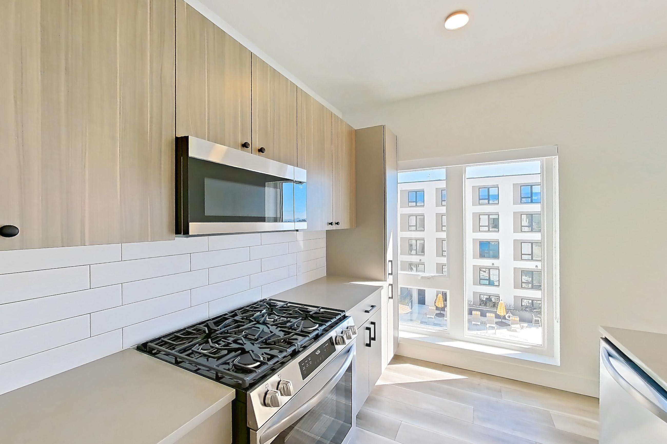 Kitchen with dishwasher, gas range, kitchen island, light countertops, light stone countertops, light wood-finish flooring, microwave, oven/range, and stainless steel appliances, Apartment 605 at The Myles