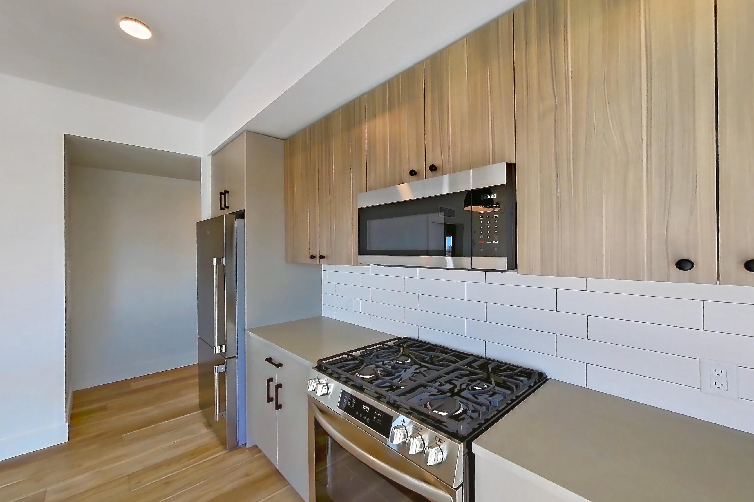 Kitchen with dishwasher, gas range, kitchen island, light countertops, light stone countertops, light wood-finish flooring, microwave, oven/range, refrigerator, stainless steel appliances, and wood-look tile flooring, Apartment 658 at The Myles