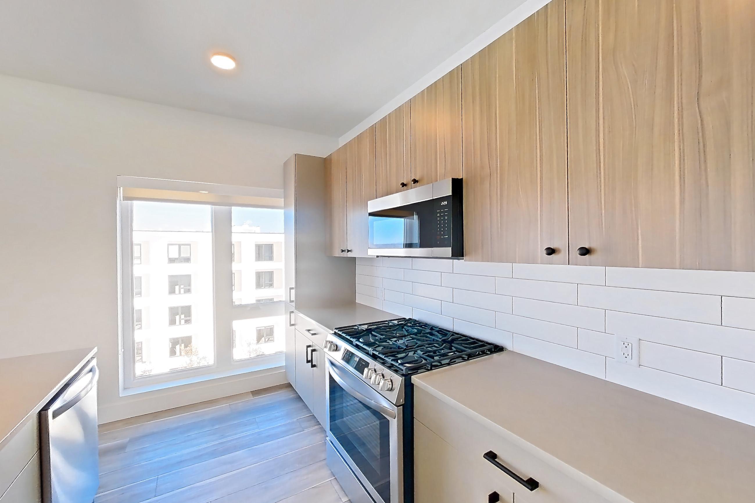 Kitchen with gas range, kitchen island, light countertops, light stone countertops, light wood-finish flooring, microwave, oven/range, and stainless steel appliances, Apartment 707 at The Myles