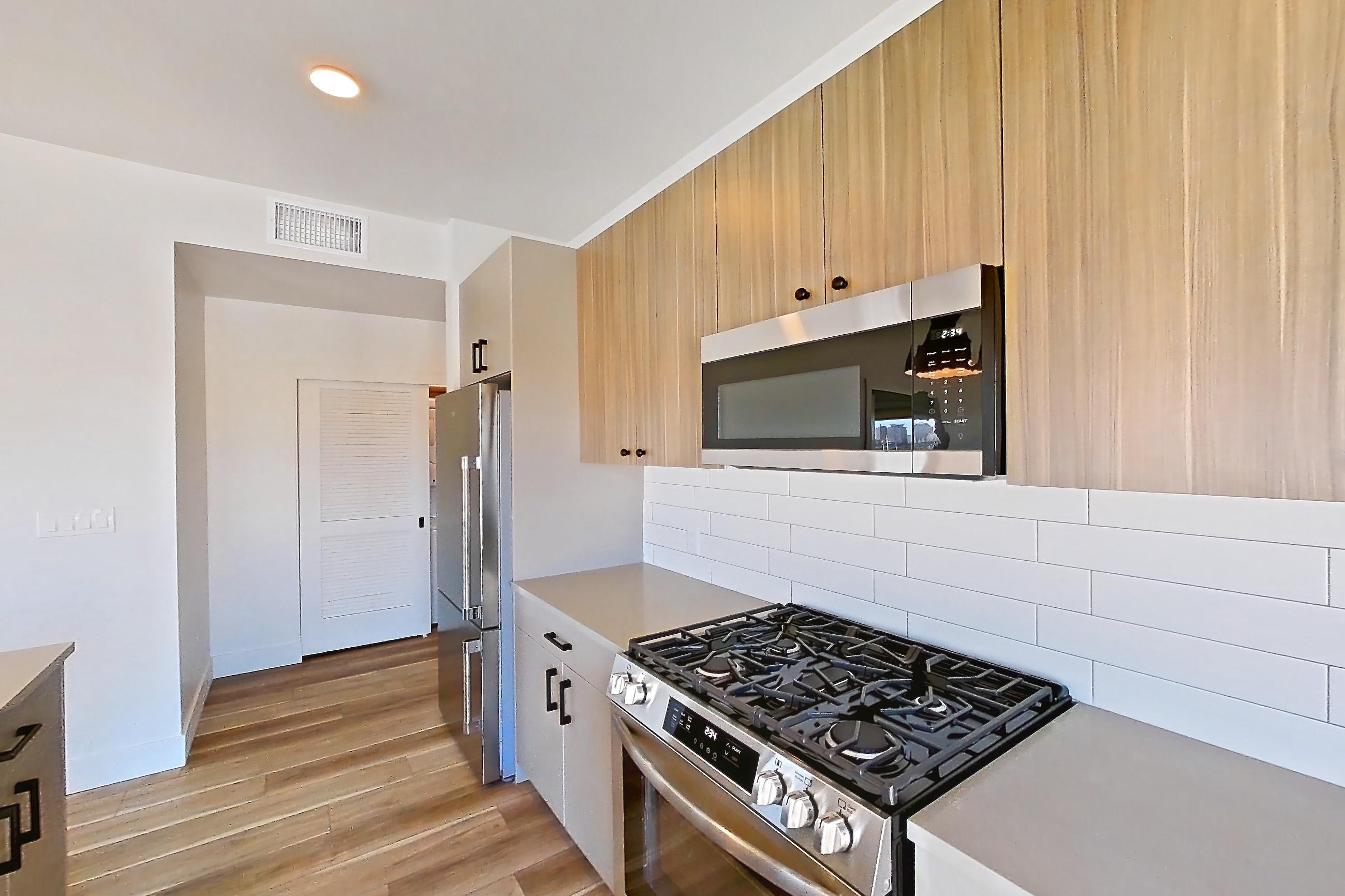 Kitchen with dark wood-finish flooring, dishwasher, gas range, kitchen island, light countertops, light stone countertops, light wood-finish flooring, oven/range, refrigerator, and stainless steel appliances, Apartment 749 at The Myles