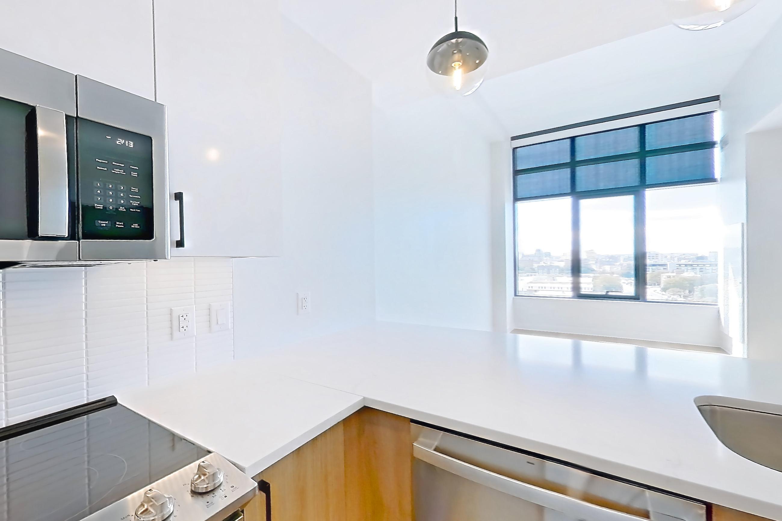 Kitchen with dishwasher, electric range, light countertops, light stone countertops, light wood-finish flooring, microwave, oven/range, refrigerator, stainless steel appliances, and water & ice dispenser, Apartment 1211 at The Alcott