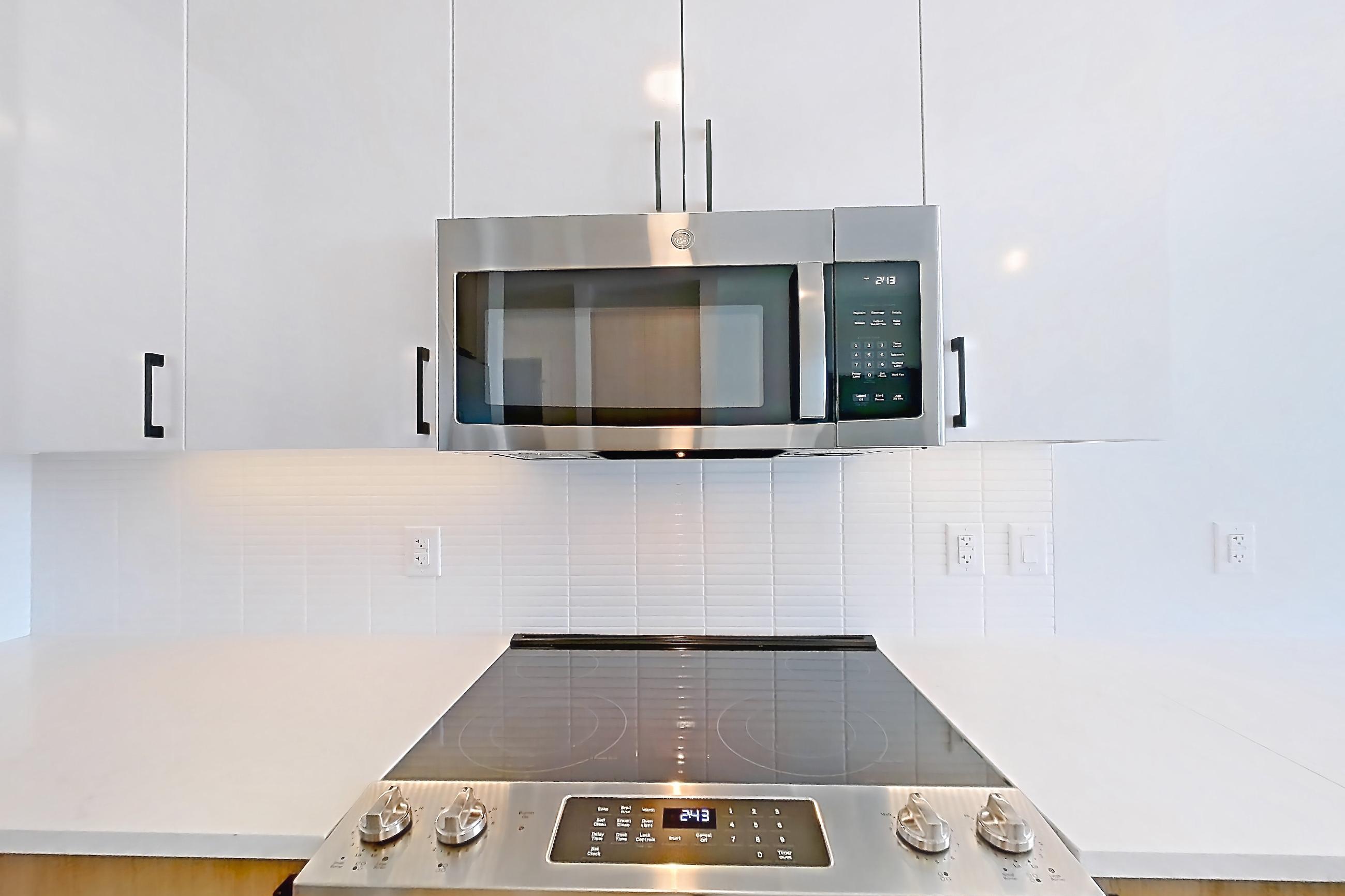 Kitchen with dishwasher, electric range, light countertops, light stone countertops, light wood-finish flooring, microwave, oven/range, refrigerator, stainless steel appliances, and water & ice dispenser, Apartment 1211 at The Alcott
