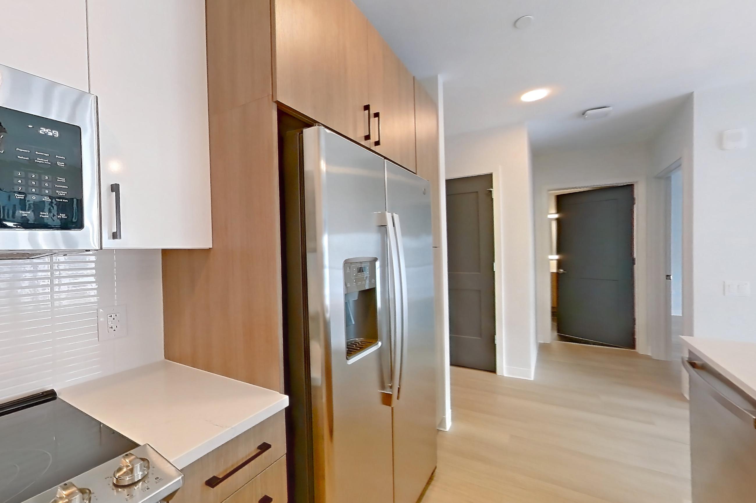 Kitchen with dishwasher, electric range, light countertops, light stone countertops, light wood-finish flooring, microwave, oven/range, refrigerator, stainless steel appliances, and water & ice dispenser, Apartment 1110 at The Alcott