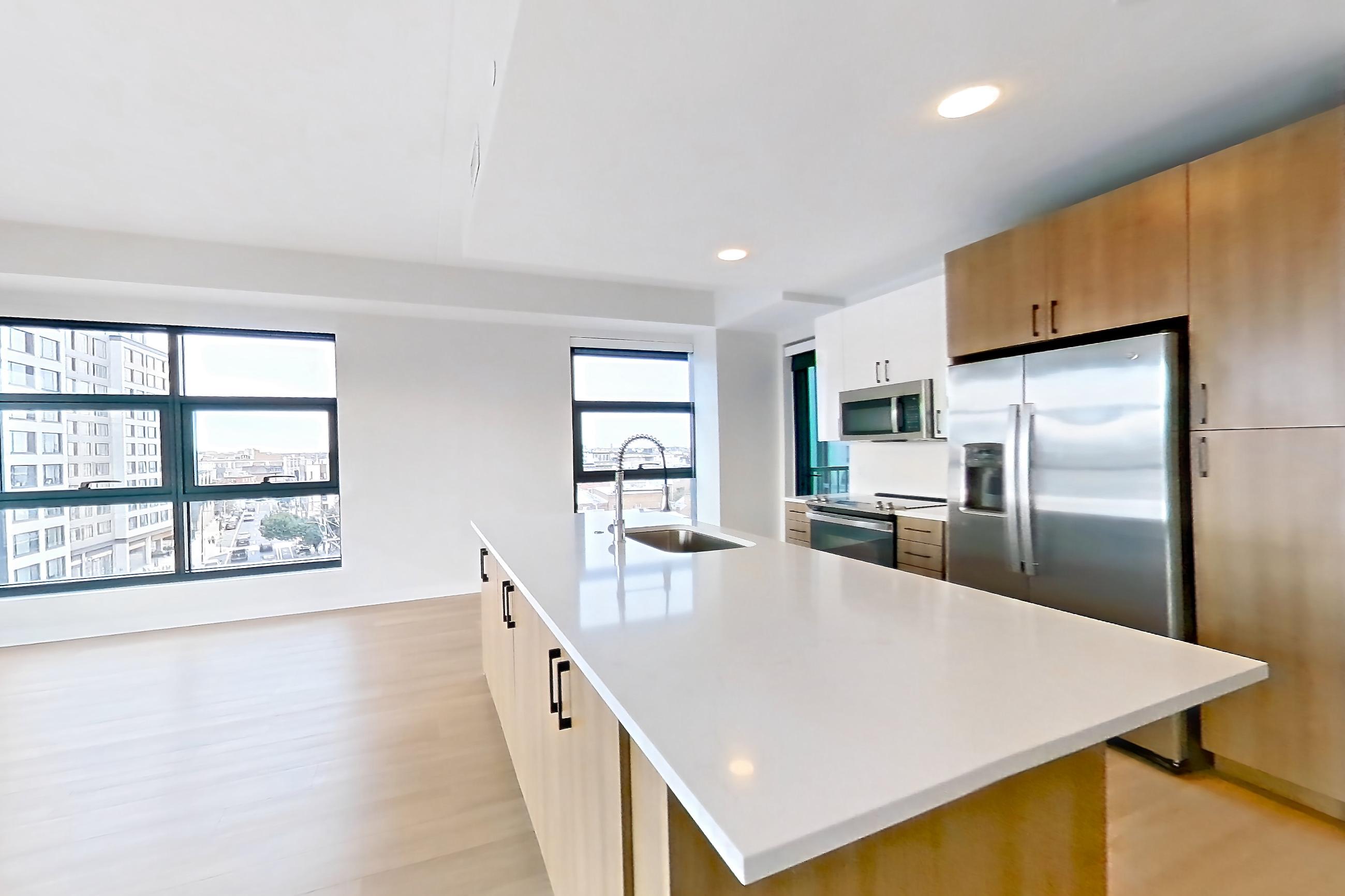Living space with electric range, kitchen island, light countertops, light stone countertops, light wood-finish flooring, microwave, oven/range, refrigerator, stainless steel appliances, and water & ice dispenser, Apartment 0615 at The Alcott